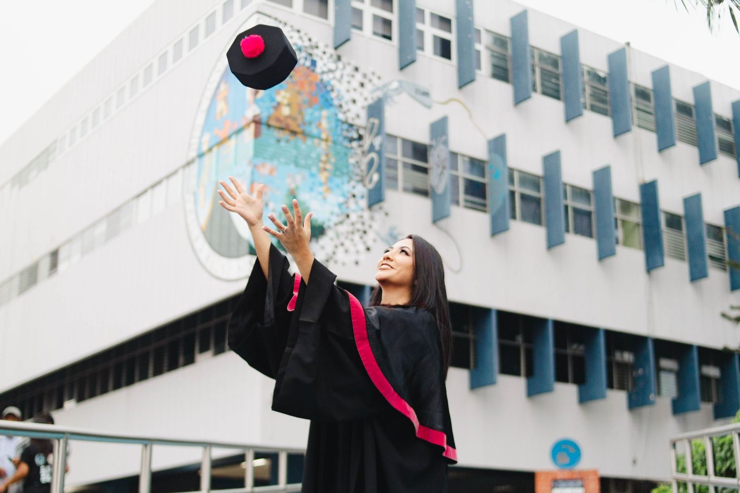 Graduate in black academic regalia tosses graduation cap in celebration outside modern building.