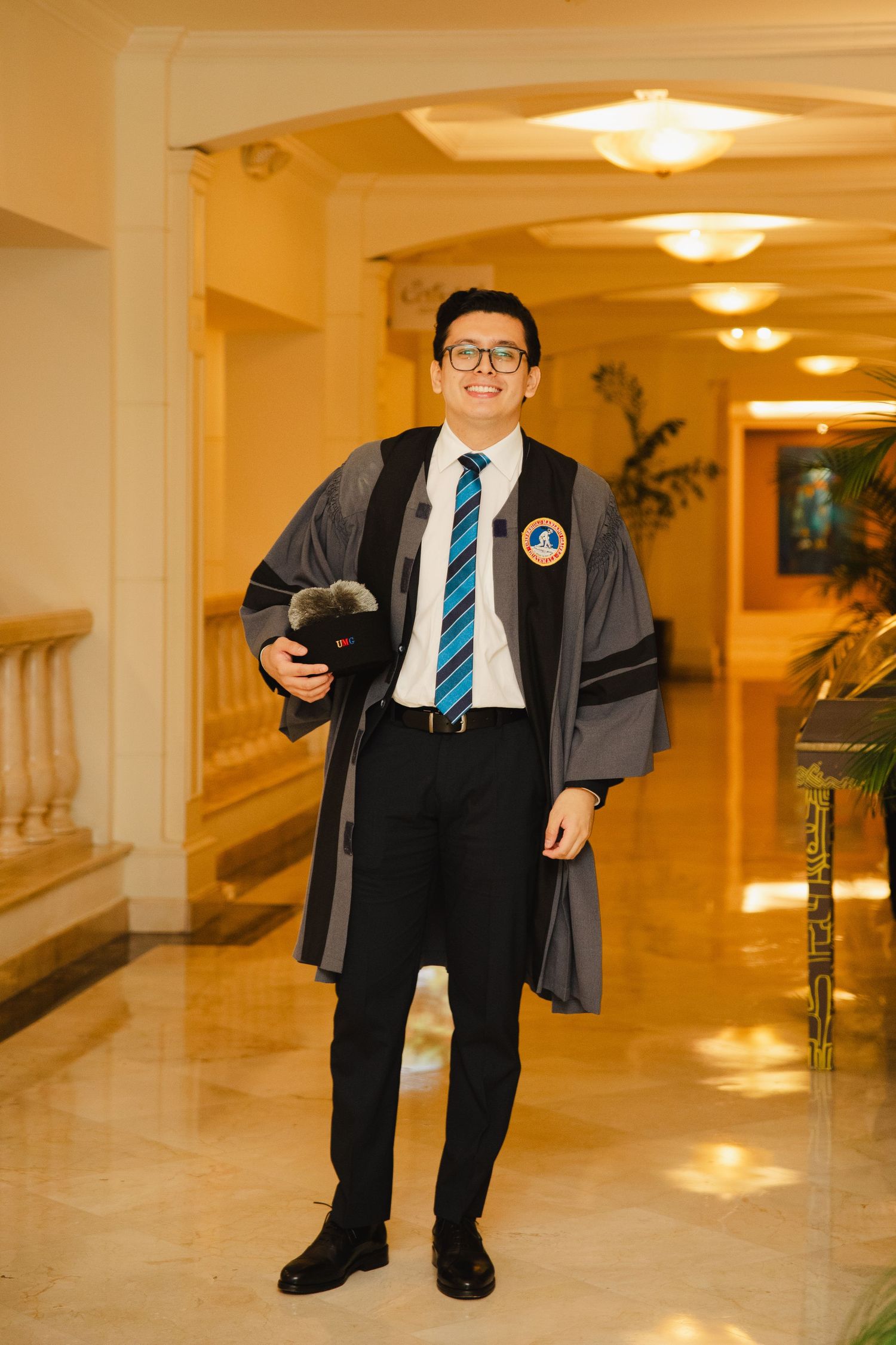 Young man in graduation gown and cap smiling in elegant hall, wearing glasses and striped tie.