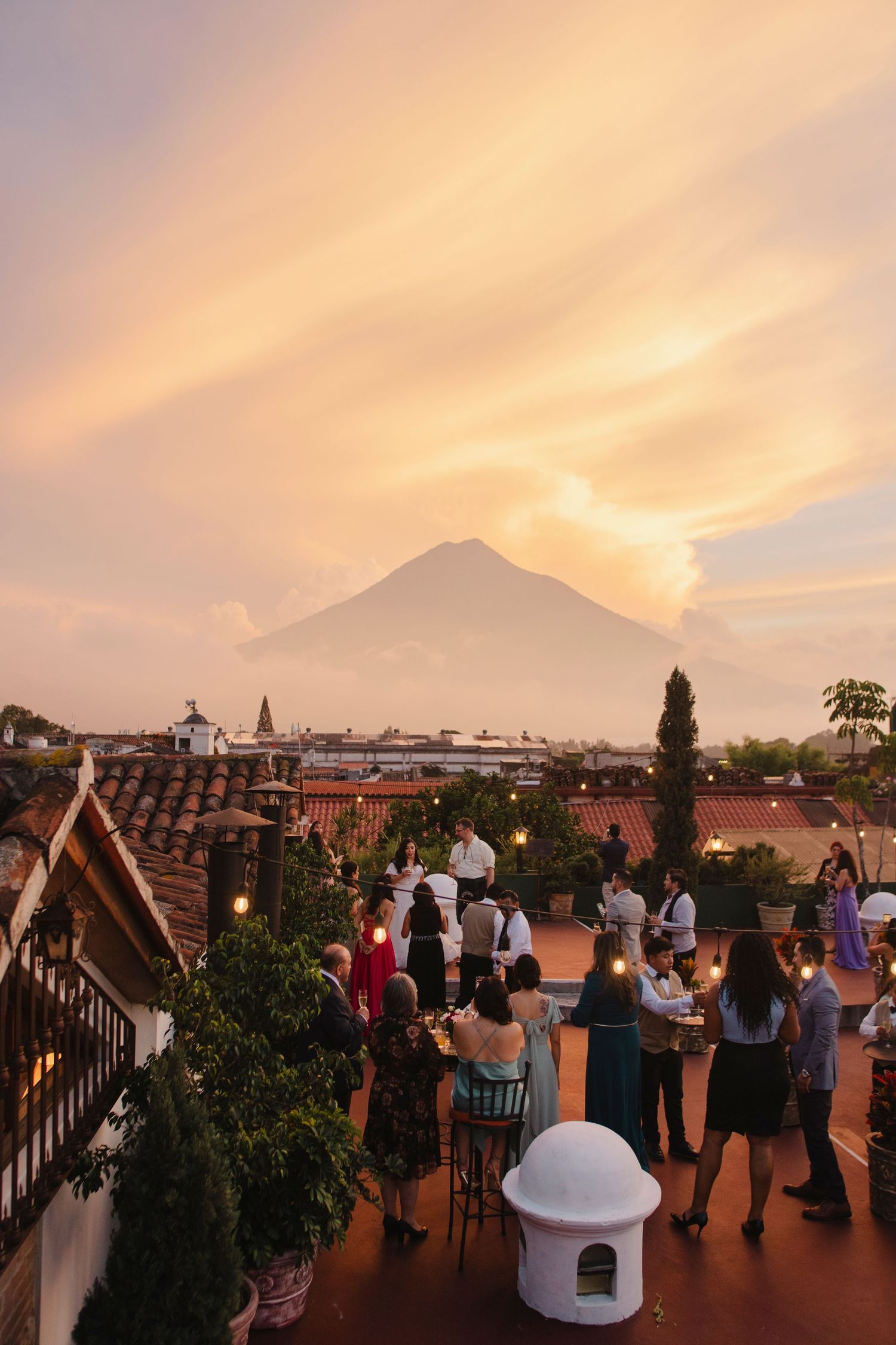 Tourists gather on a rooftop terrace at sunset with a dramatic view of a volcano silhouetted against an orange sky.