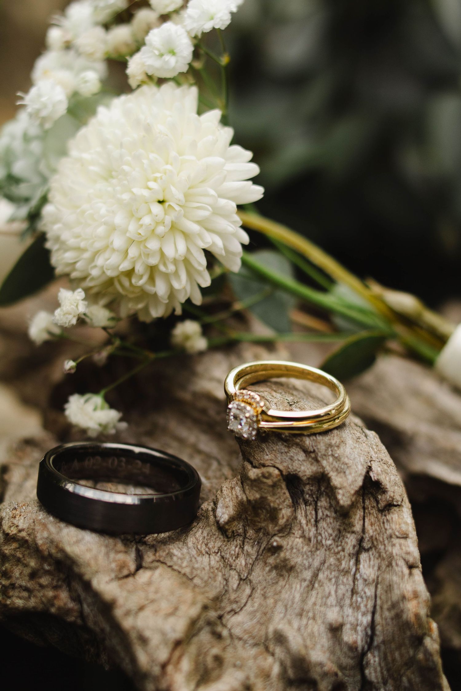 Wedding rings and white chrysanthemum flower displayed on rustic driftwood in an artistic arrangement.