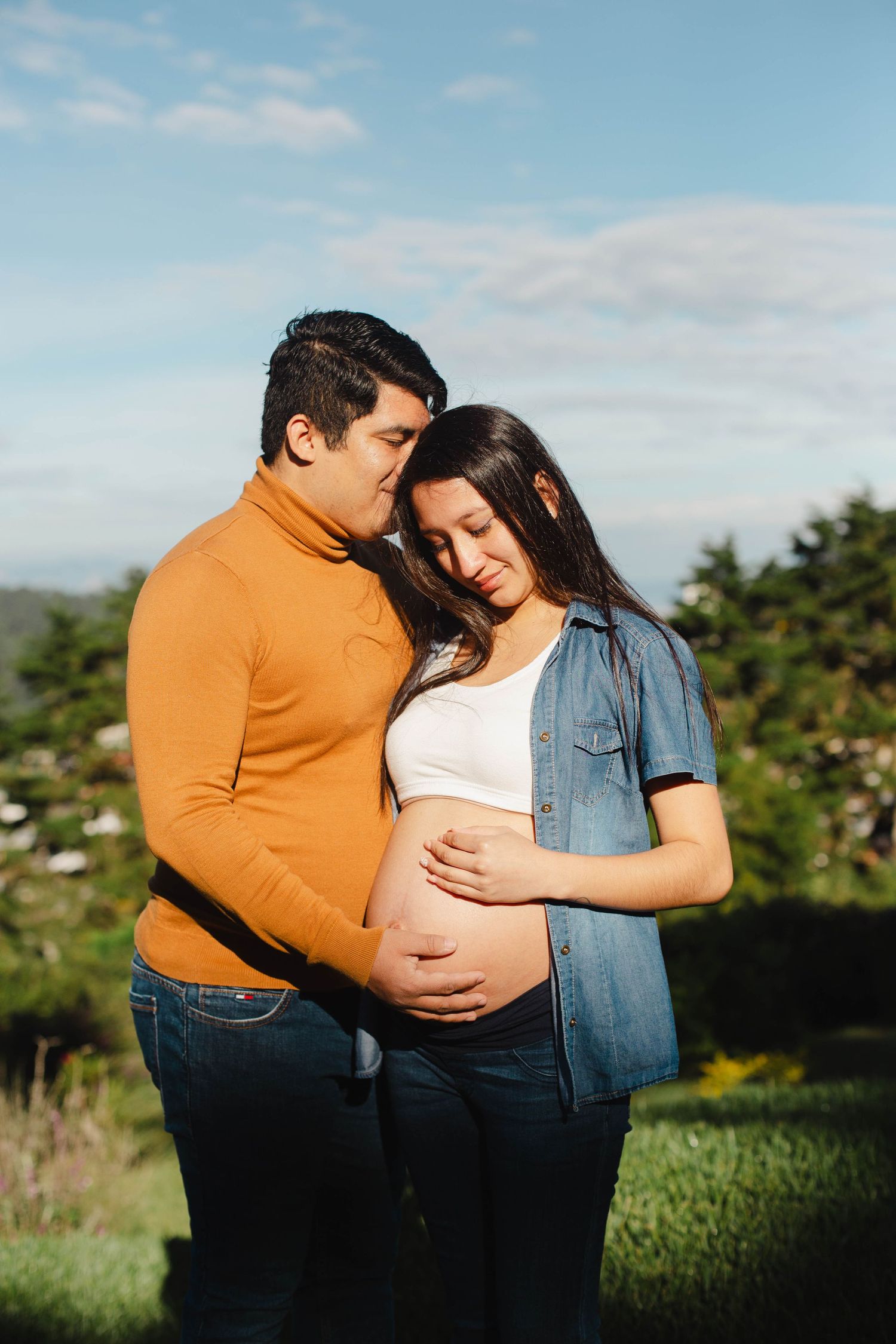 Expecting couple embracing outdoors against blue sky and mountain backdrop.