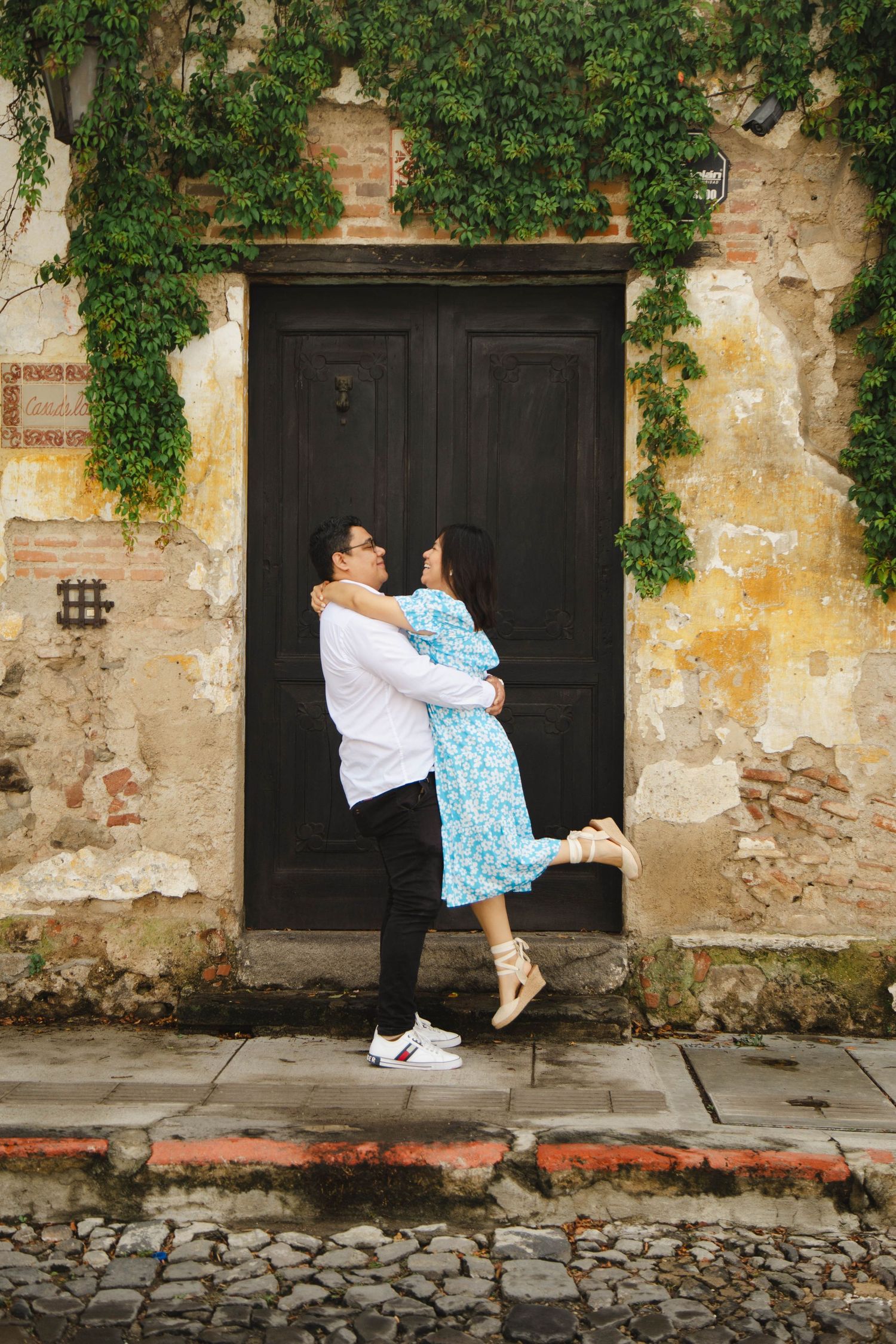 Playful couple posing by ivy-covered wall with black wooden door in colonial setting.