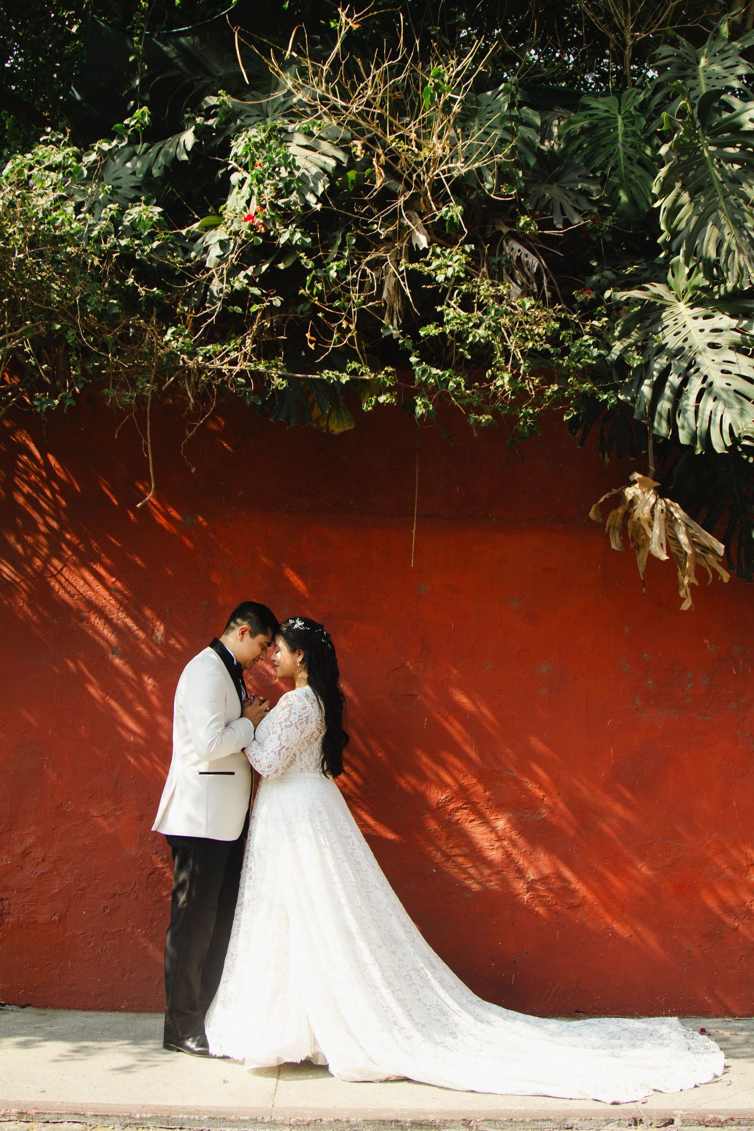 Couple in wedding attire share an intimate moment against a dramatic red wall with overhanging foliage.