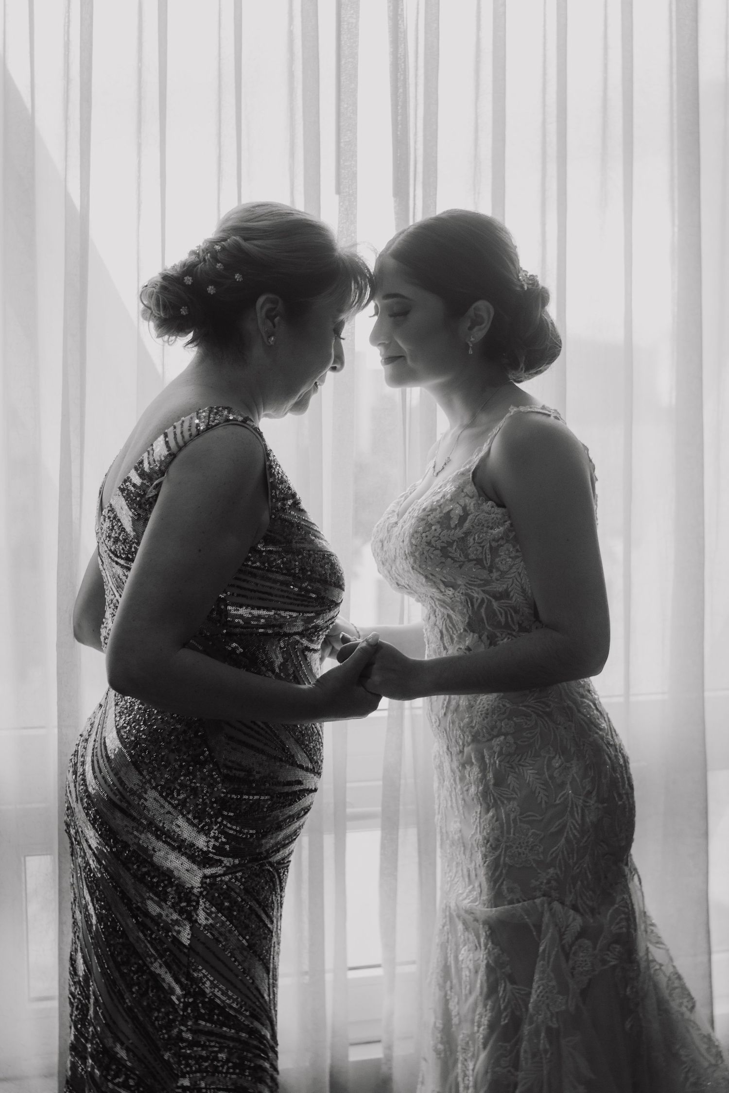 Two silhouetted figures in formal dresses share an intimate moment against sheer curtains.
