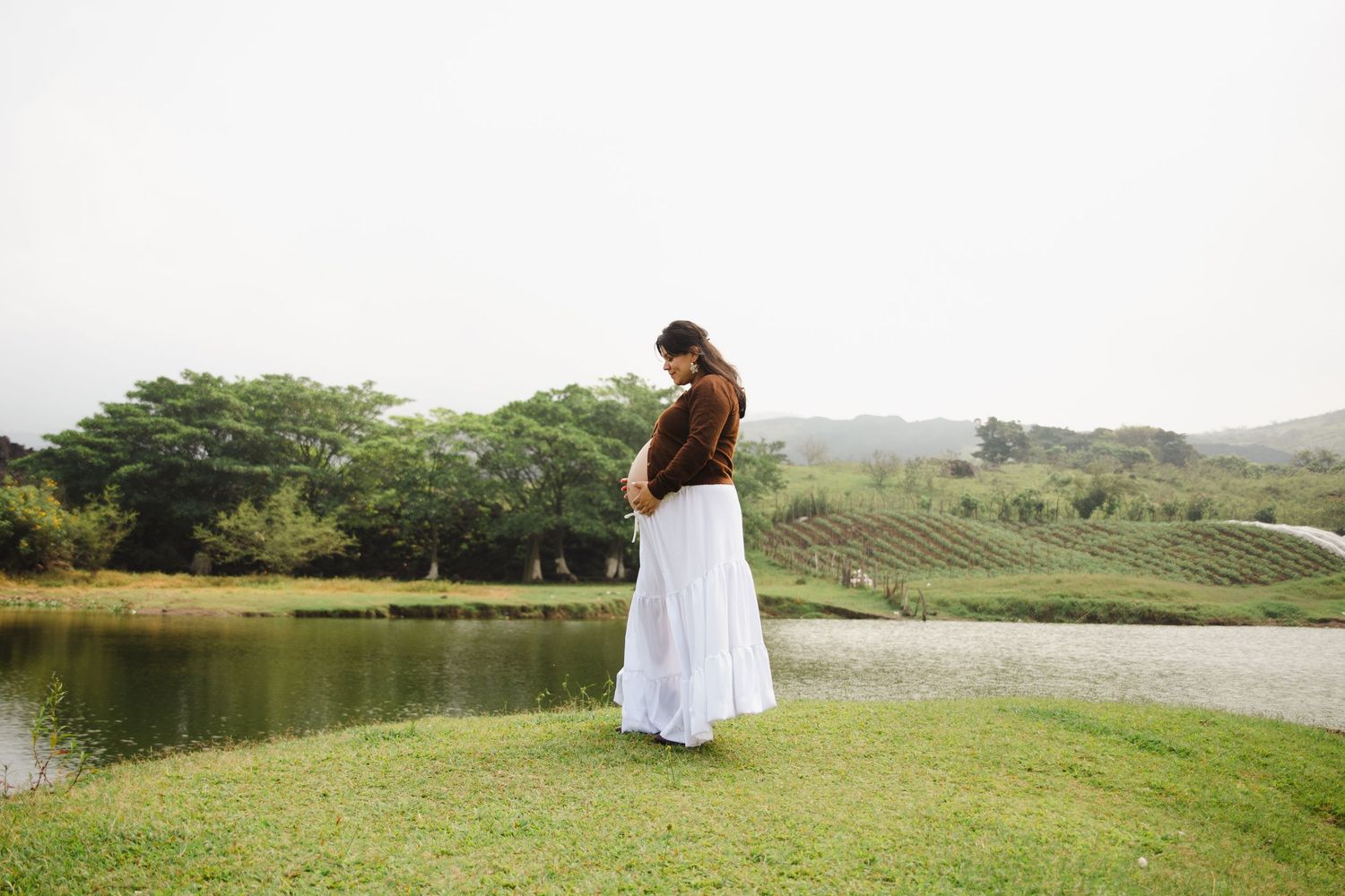 Pregnant woman in white skirt and brown top standing by a serene pond surrounded by lush greenery.