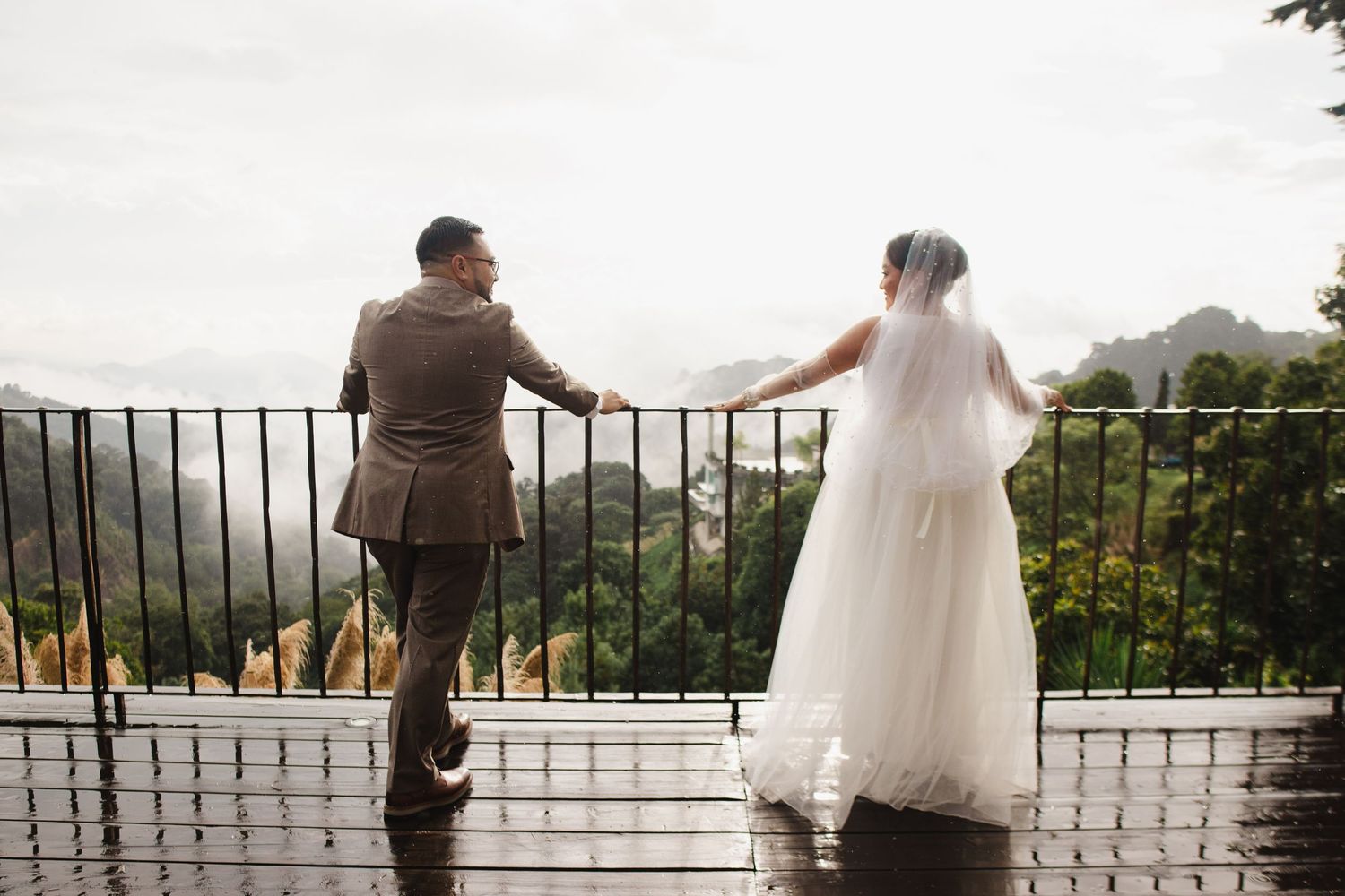 A couple in wedding attire stands at a scenic overlook with mountains and trees in the background.