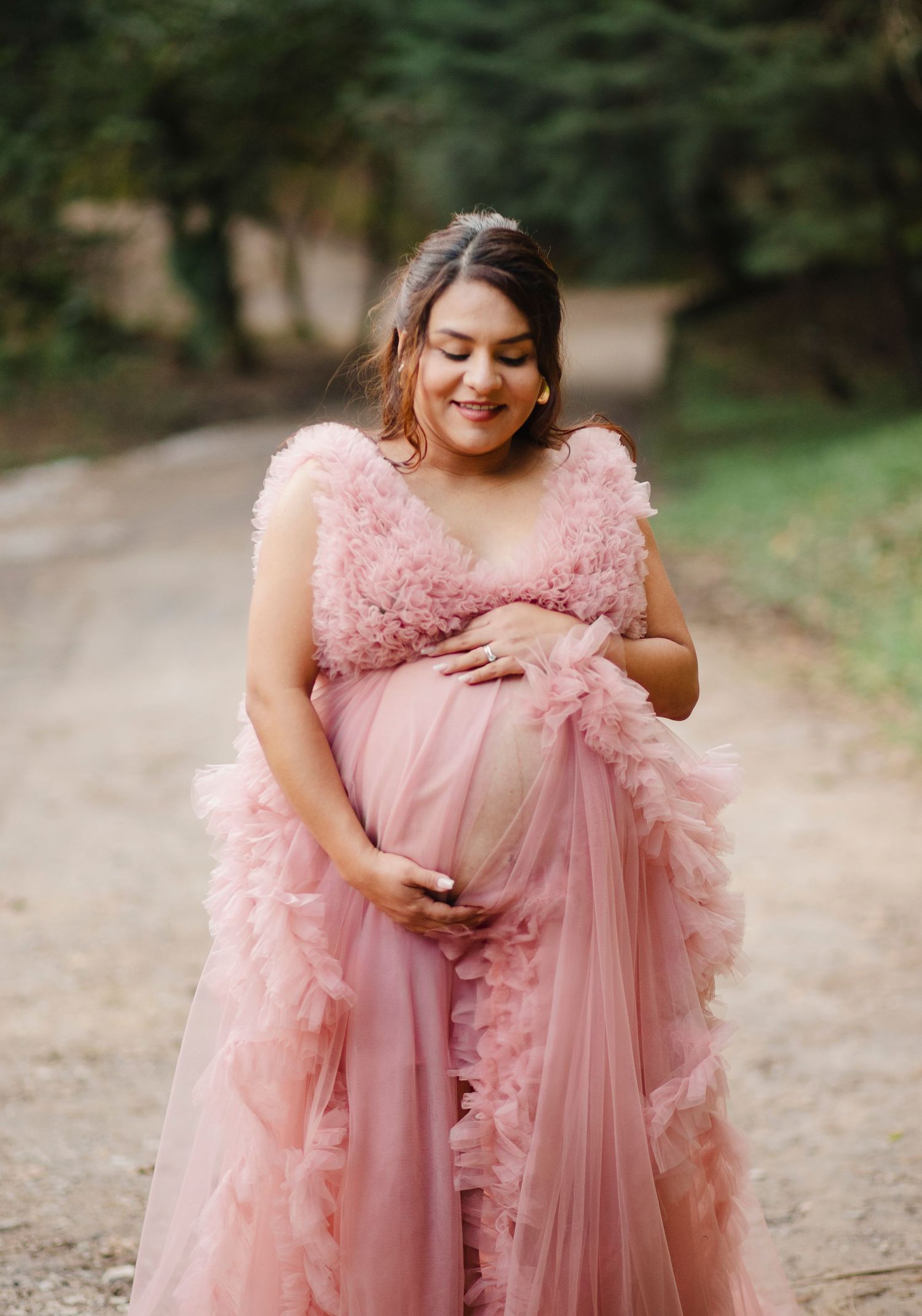 Expectant mother in flowing pink tulle maternity gown poses joyfully on a dirt path surrounded by greenery.