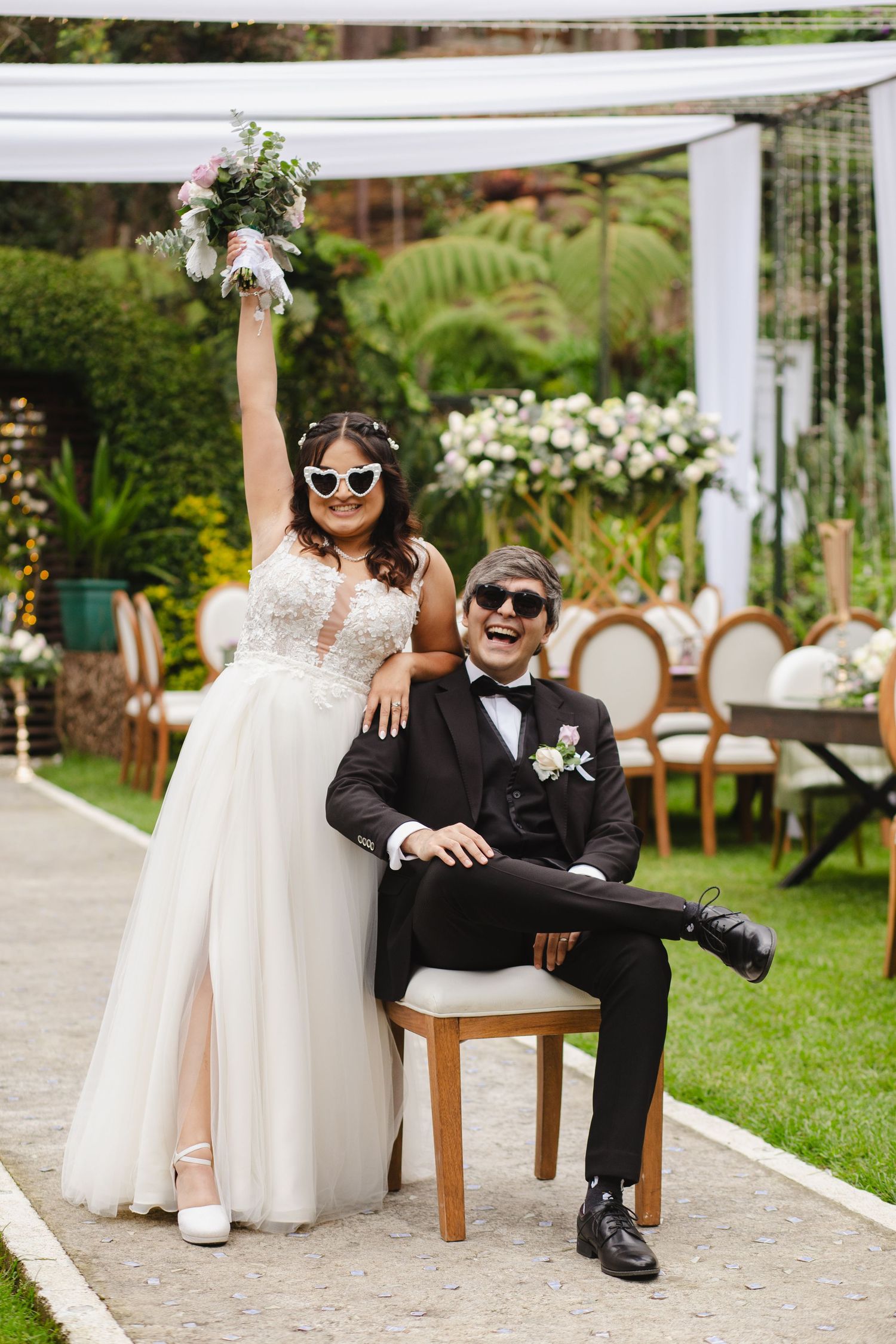 A playful outdoor garden wedding scene with sunglasses and floral decor under a white pergola.