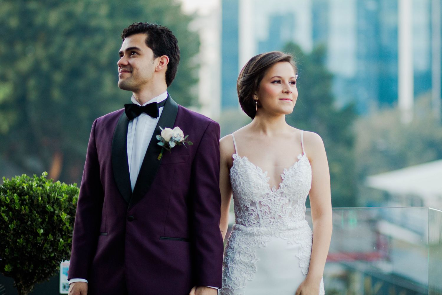 Elegant couple in purple tuxedo and white dress pose against city backdrop.
