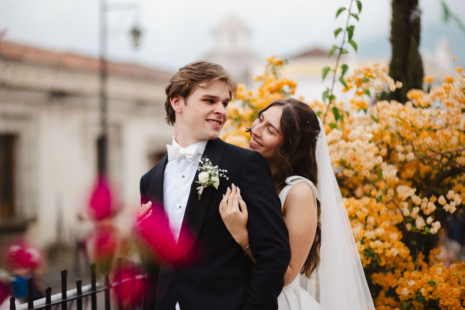 Newlywed couple embraces outdoors with beautiful orange autumn foliage and classic architecture in background.