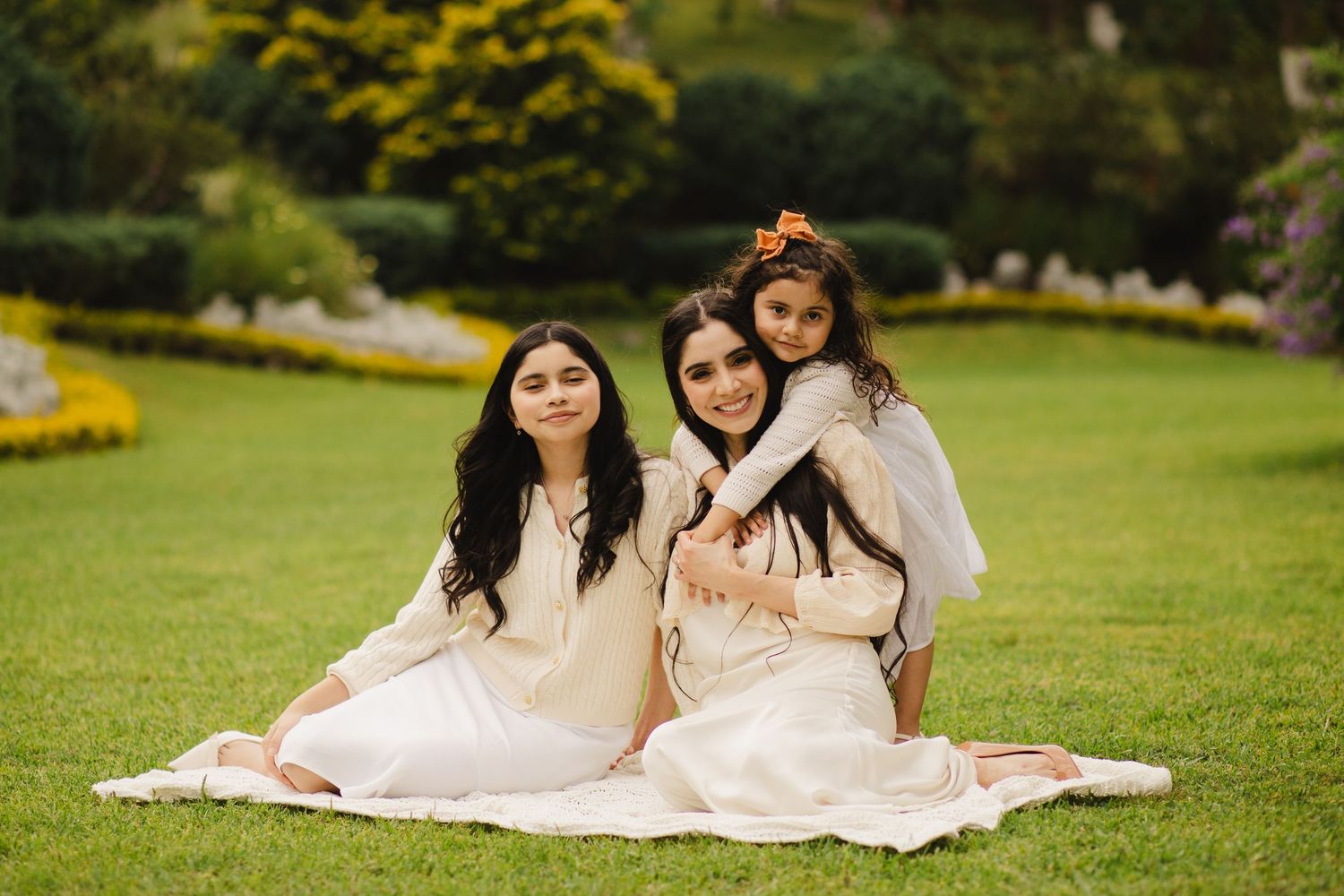 Three girls in matching white outfits sitting on grass in a lush green garden, smiling warmly for a family portrait.