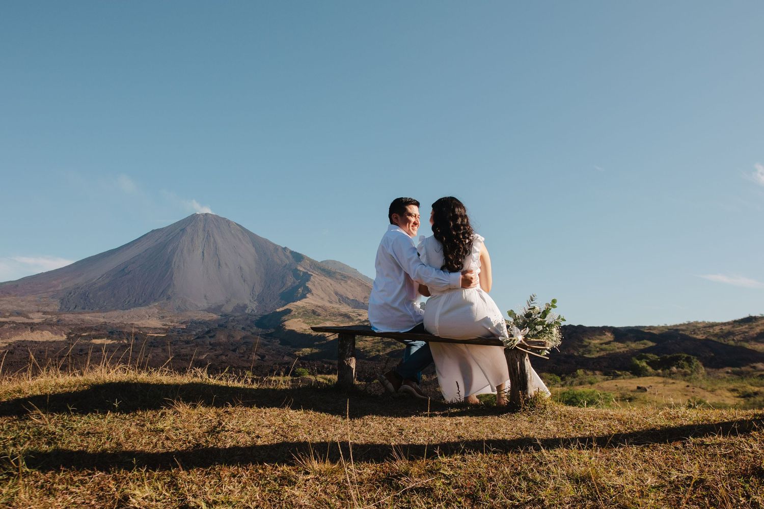 A couple in wedding attire sits together overlooking a scenic mountain landscape during sunset.