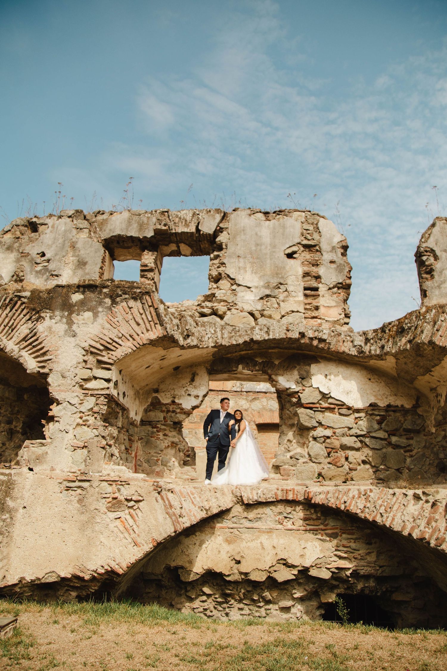 A couple in wedding attire poses in an archway of historic stone ruins.