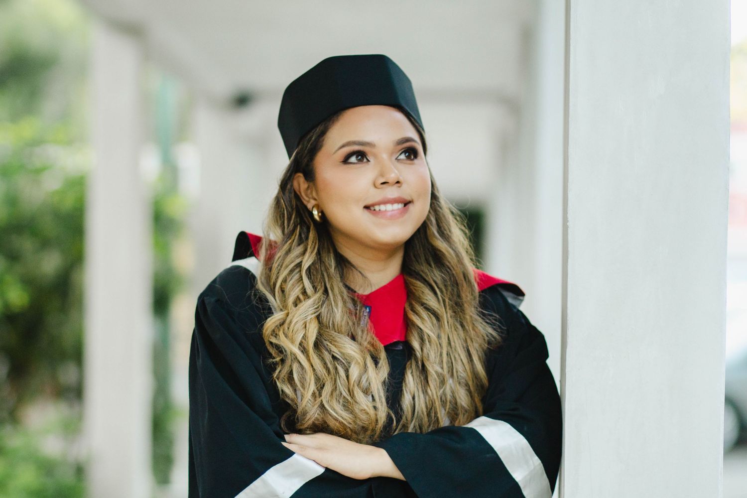 Graduate wearing academic regalia and red shirt smiles while posing against white column.