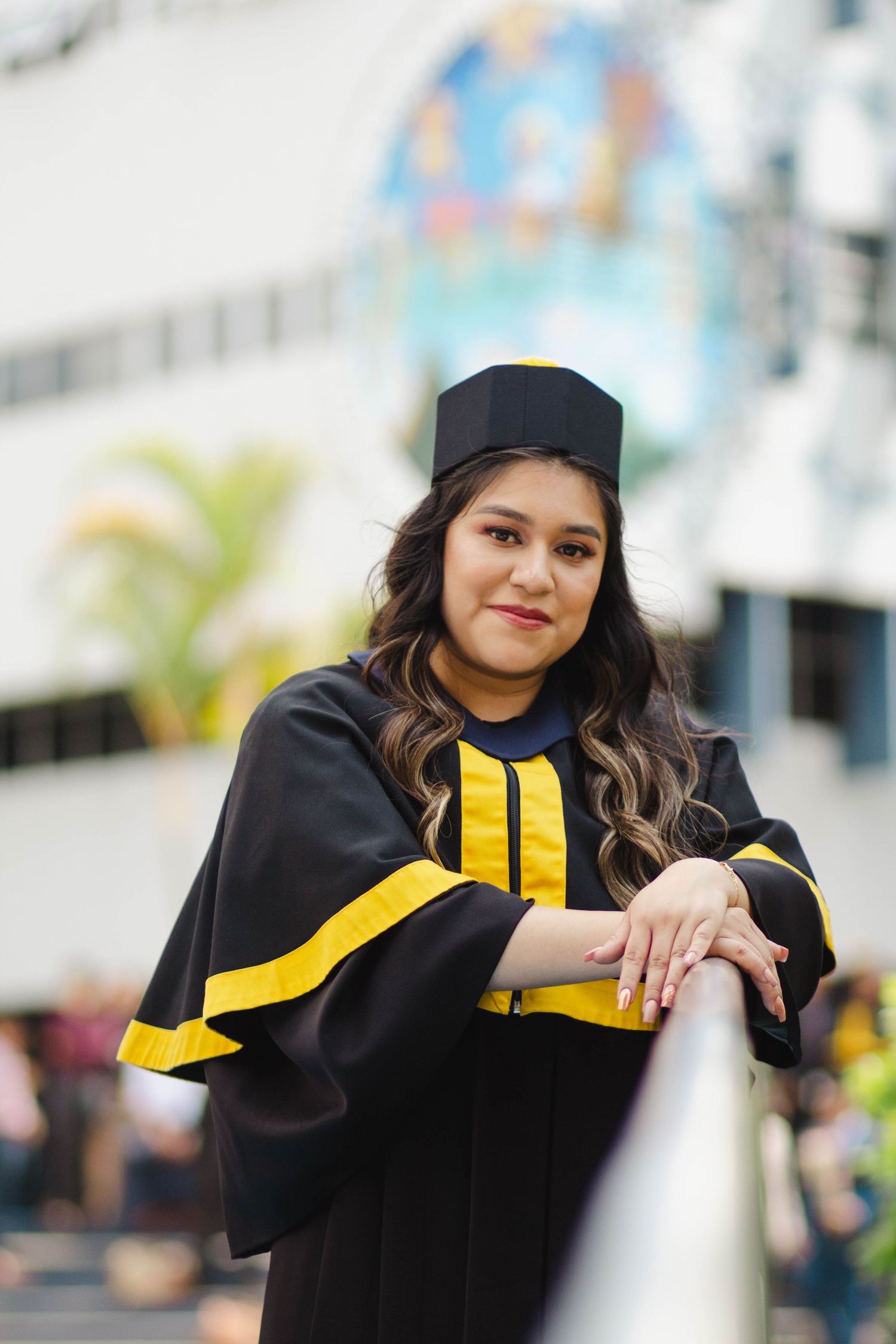 Graduate in black and yellow academic regalia poses against wall with urban mural background.