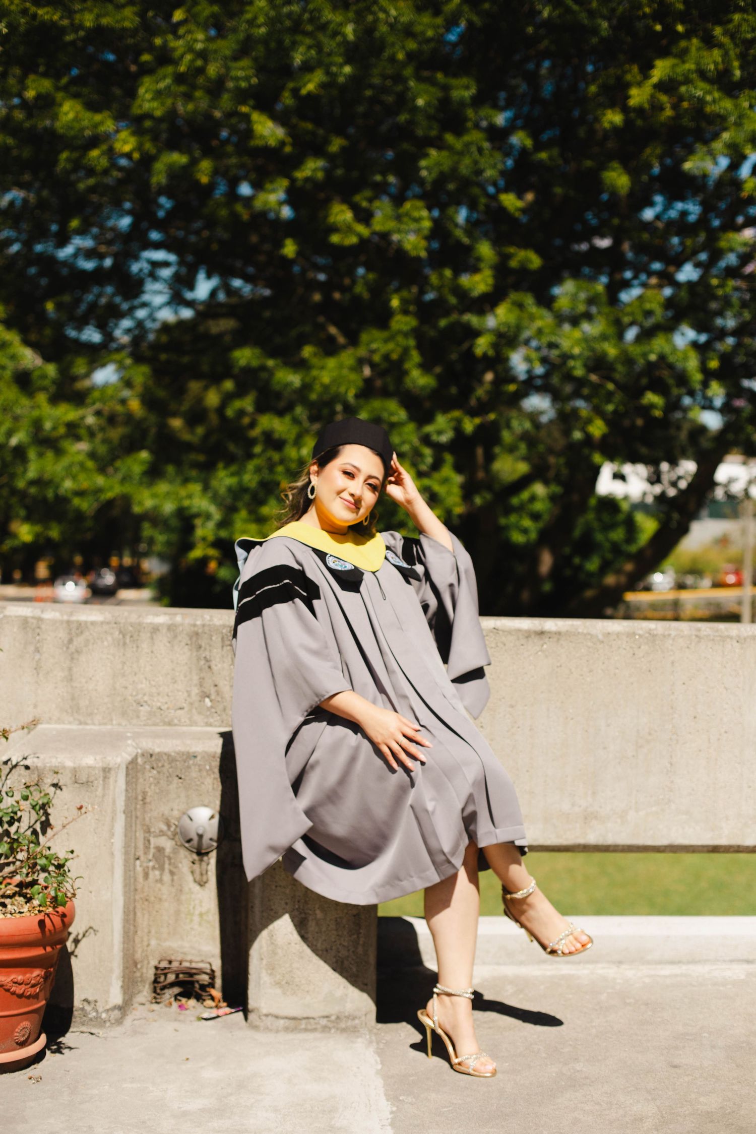 Graduate in grey and yellow academic regalia poses casually on concrete ledge outdoors.