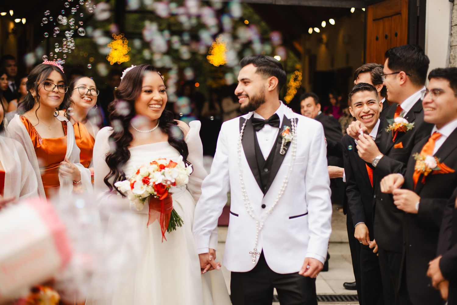 Wedding party celebrates with couple in white attire walking through venue.