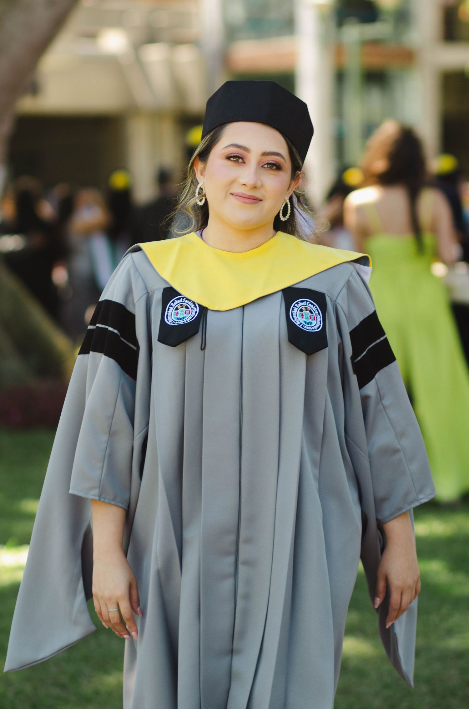 Graduate poses in grey academic gown with yellow stole and mortarboard cap on campus lawn.