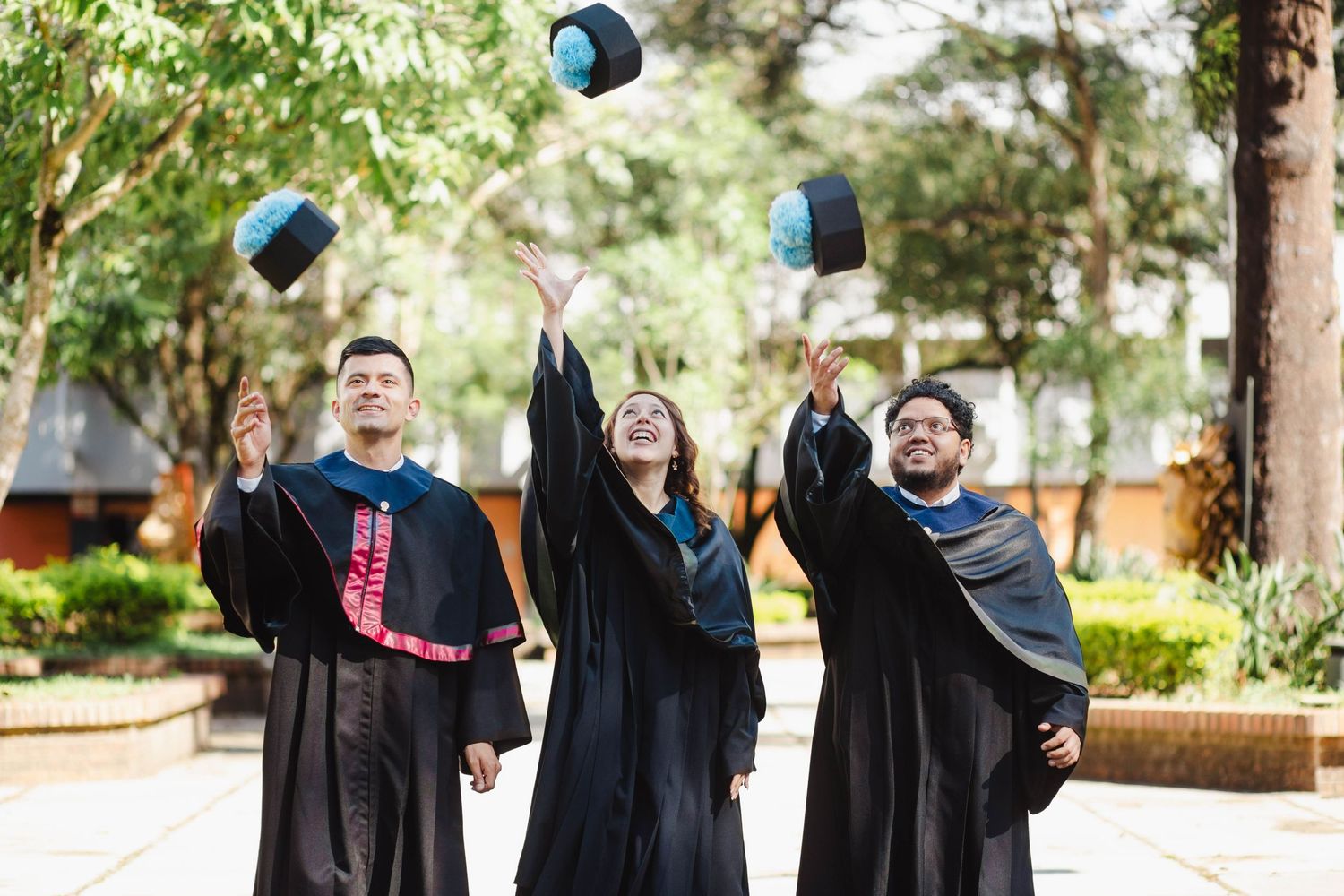 Three graduates in black academic regalia toss their caps in celebration outdoors.