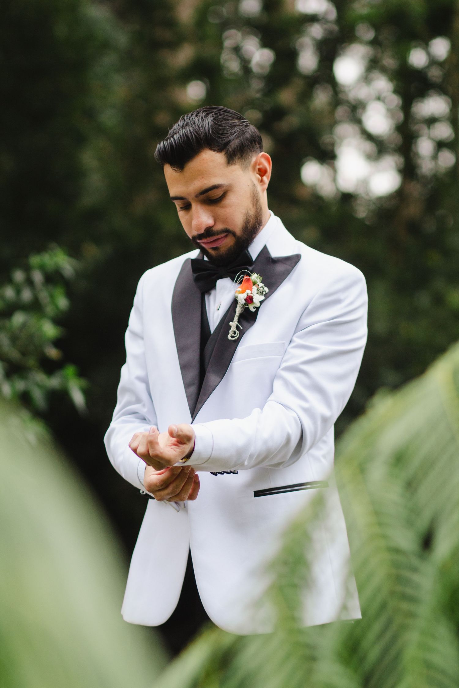 Groom in white tuxedo jacket adjusts cufflink while standing among tropical foliage.