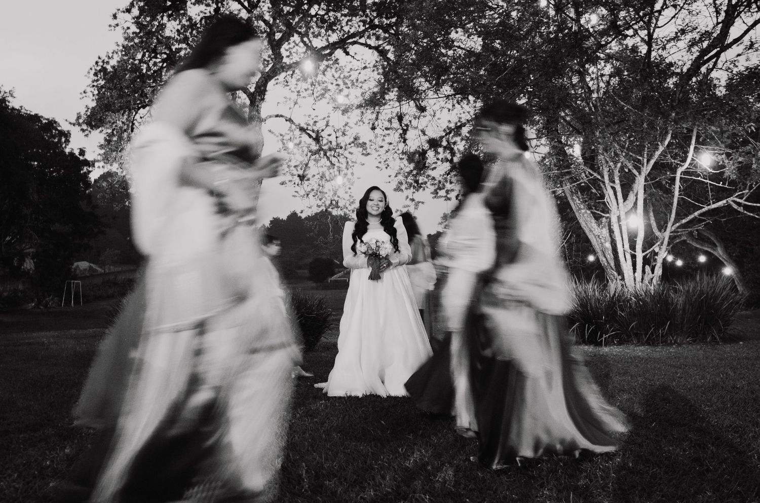 Motion-blurred black and white wedding scene in a garden setting with figures dancing around a seated bride.