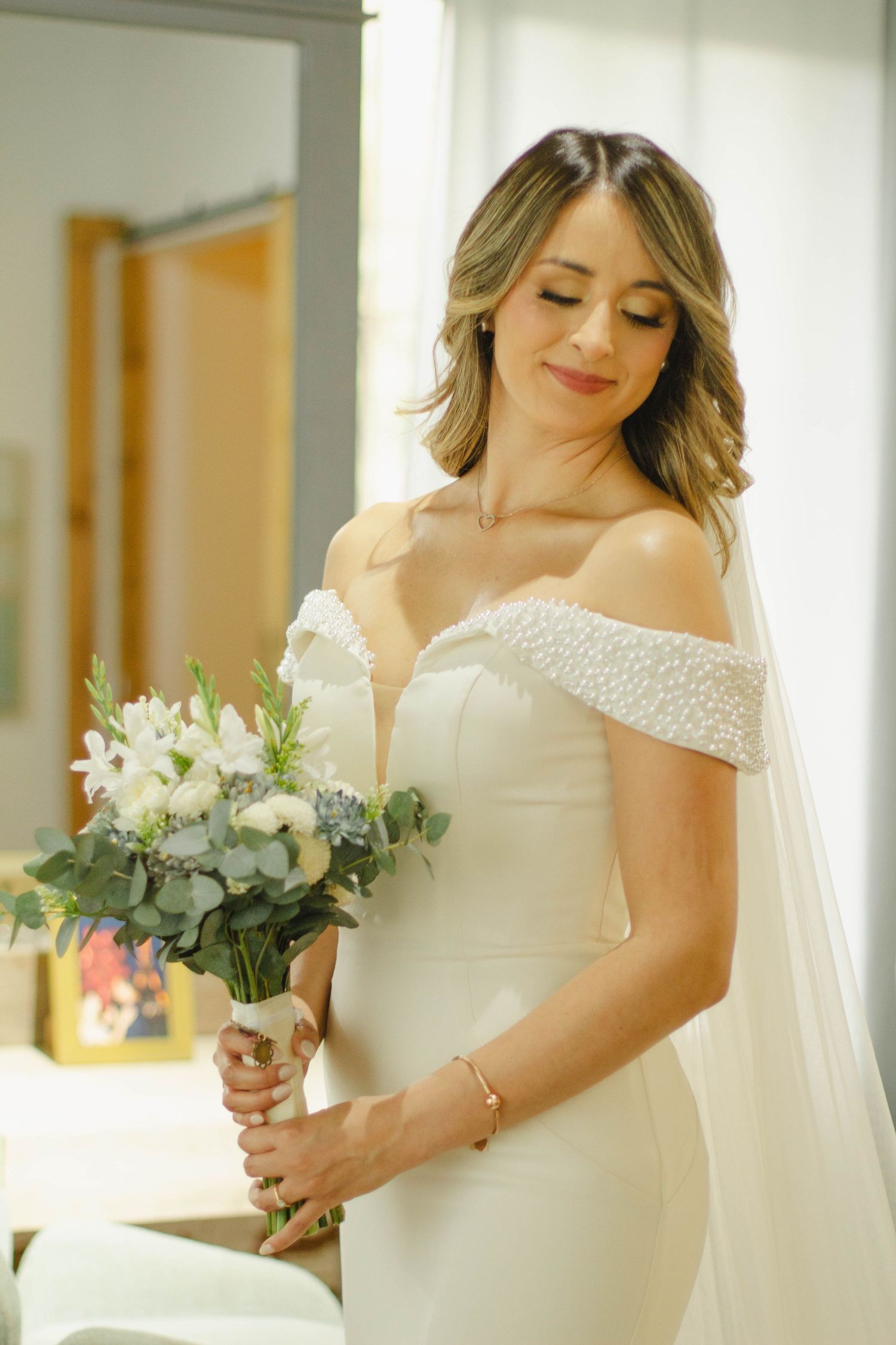 Elegant bride in off-shoulder white dress holding delicate white and green bouquet.