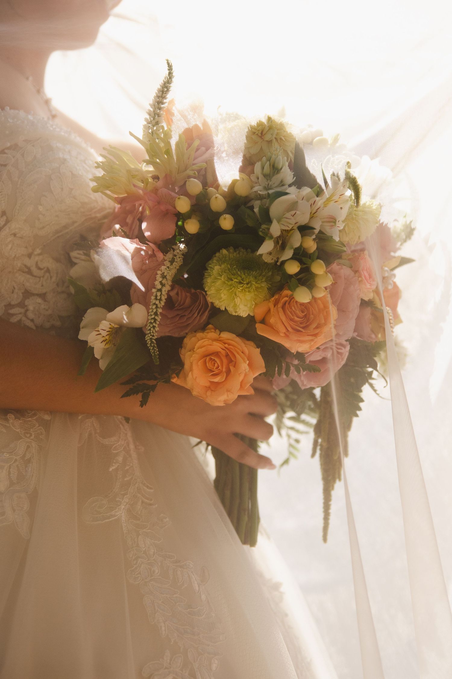 Bride in white lace gown holding a romantic bouquet of peach roses, green blooms, and mixed flowers in soft natural light.