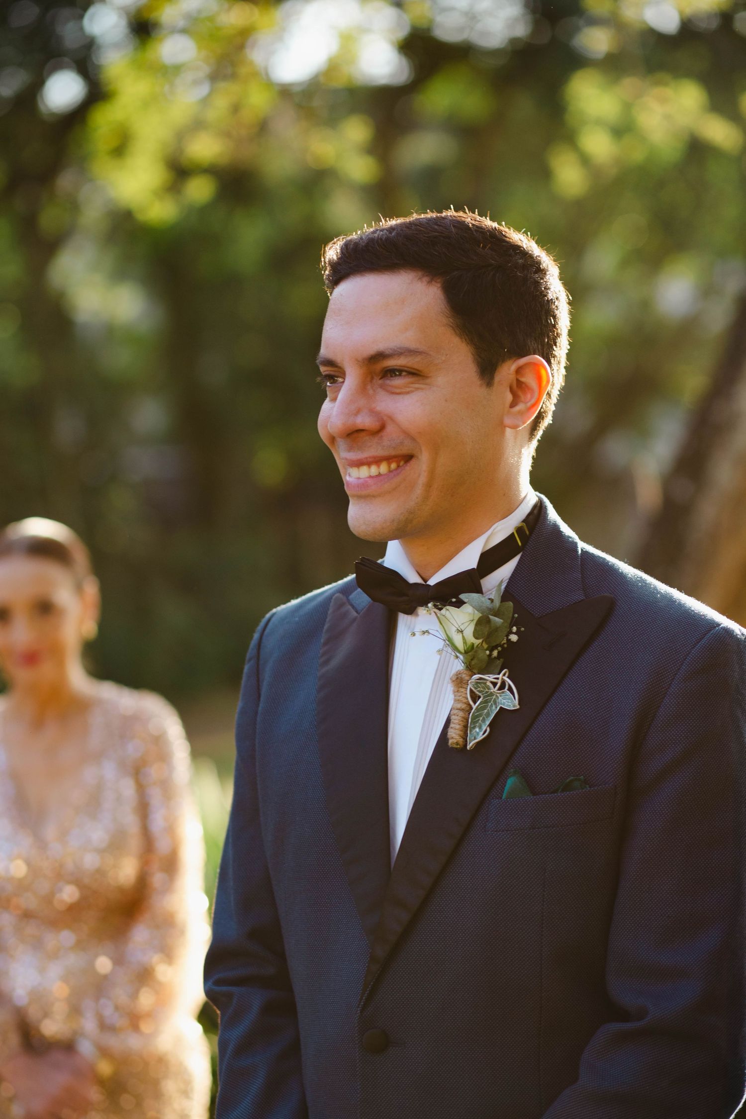 A groom in a navy tuxedo and black bow tie smiles warmly at a sunny outdoor wedding ceremony.