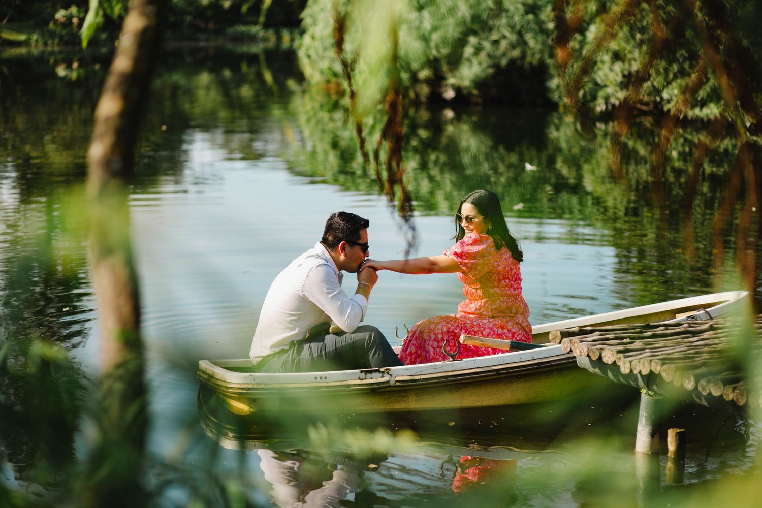A couple shares a romantic moment while sitting in a small boat on a tranquil lake surrounded by trees.