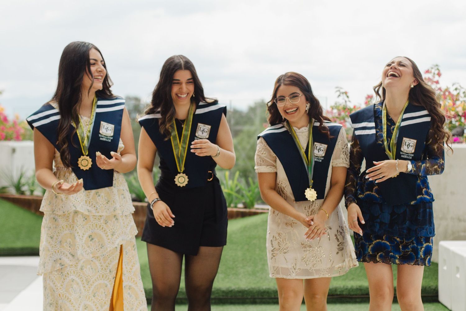 Four women in formal dresses wearing medals and lanyards laugh together at an outdoor event.