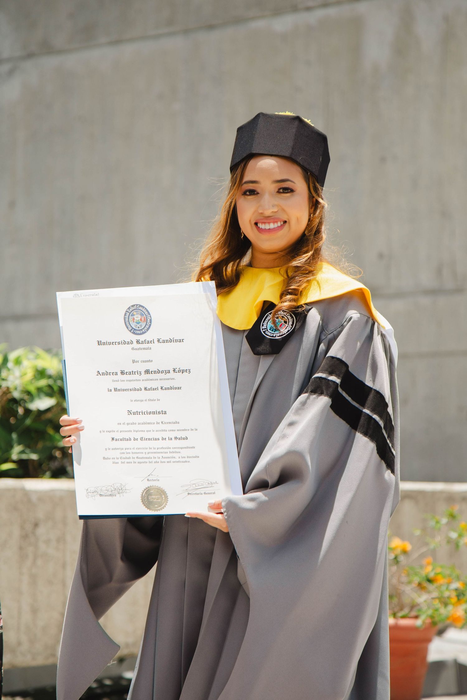 Graduate holds diploma while wearing grey academic regalia against concrete wall background.