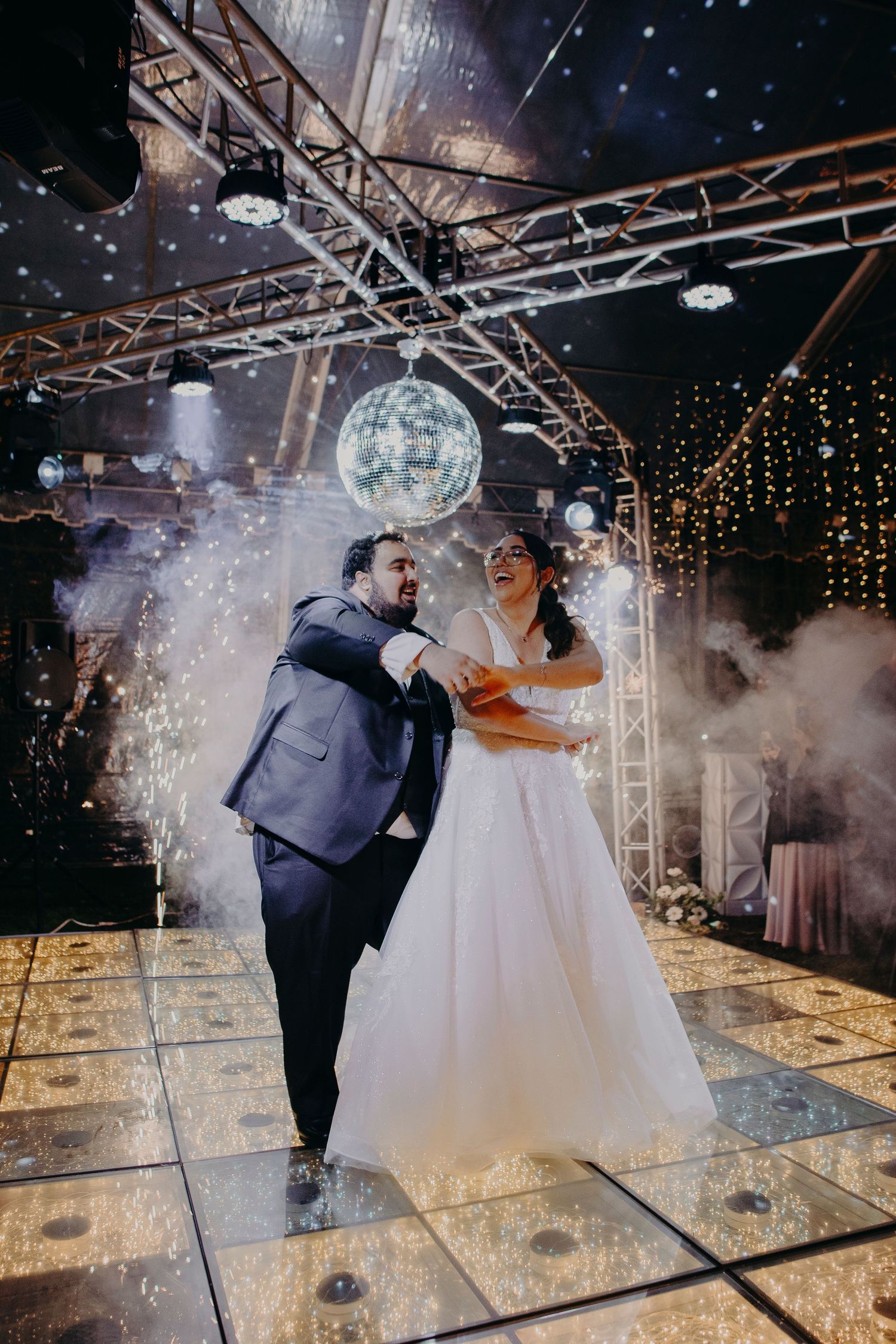 A couple shares their first dance at a wedding reception under sparkling lights and fog effects on a reflective dance floor.