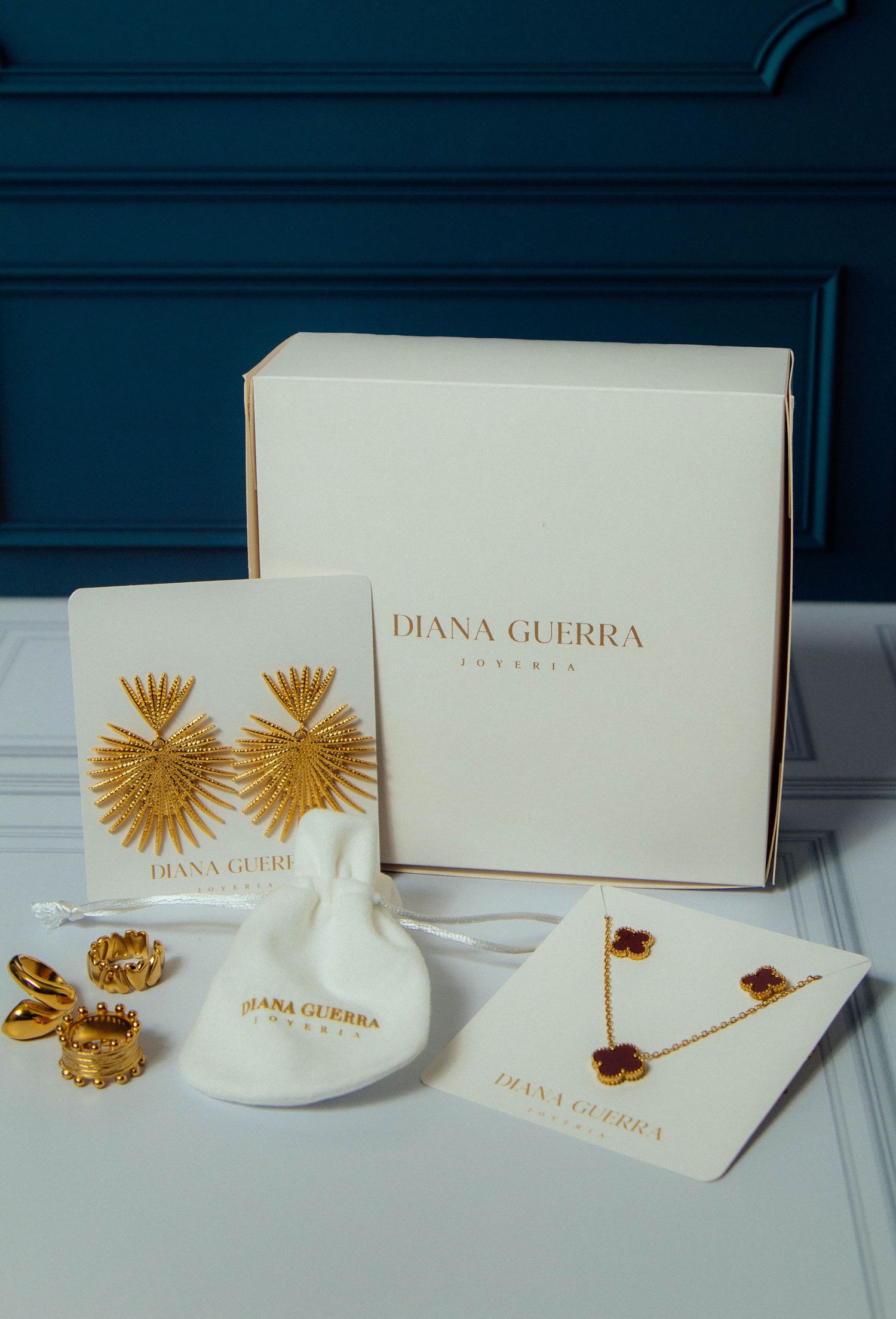 Gold tone Diana Guerra jewelry collection including starburst earrings, necklace and rings displayed with branded packaging.