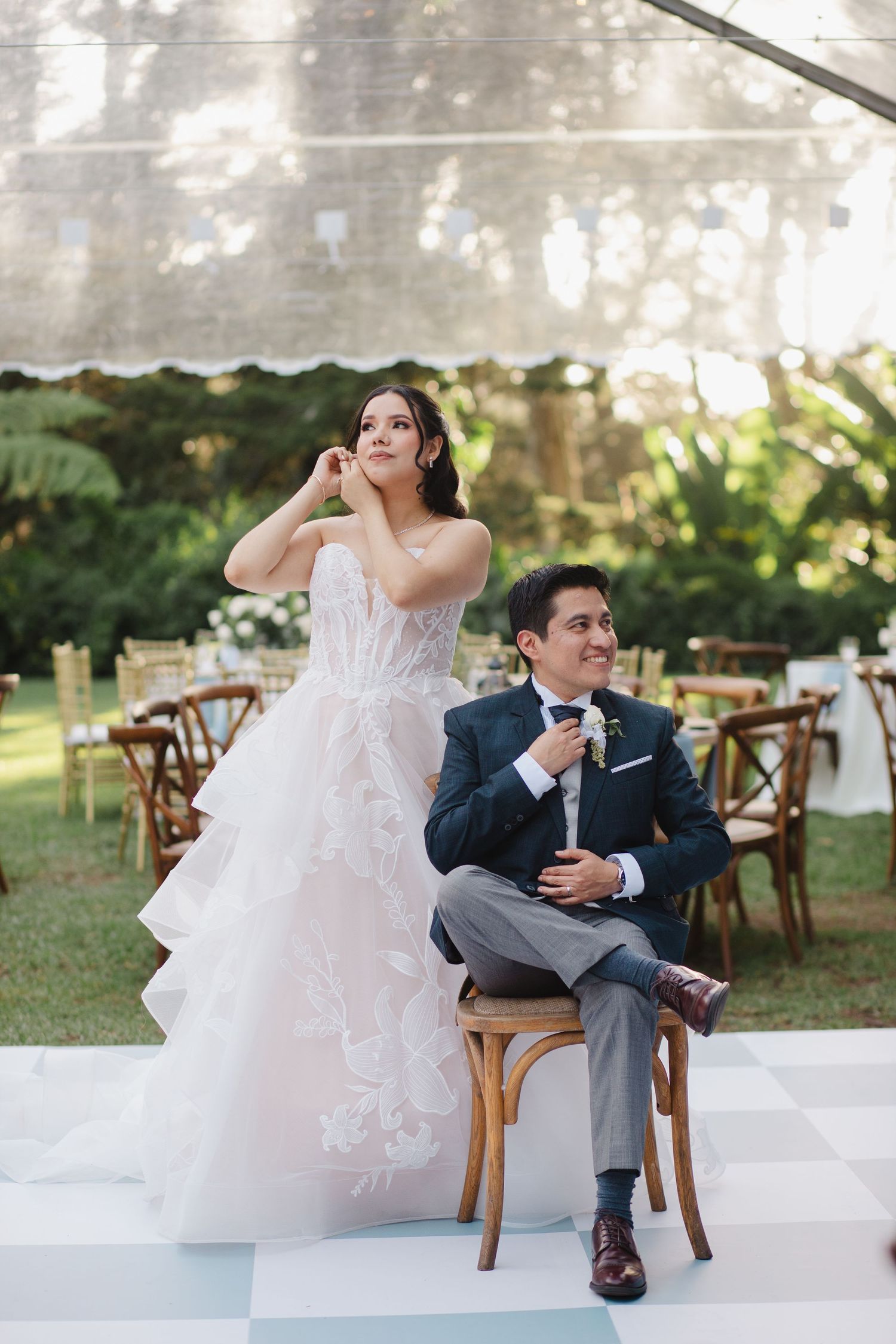 Bride in ruffled white gown and groom in navy suit sit on wooden chairs at an elegant outdoor wedding reception.