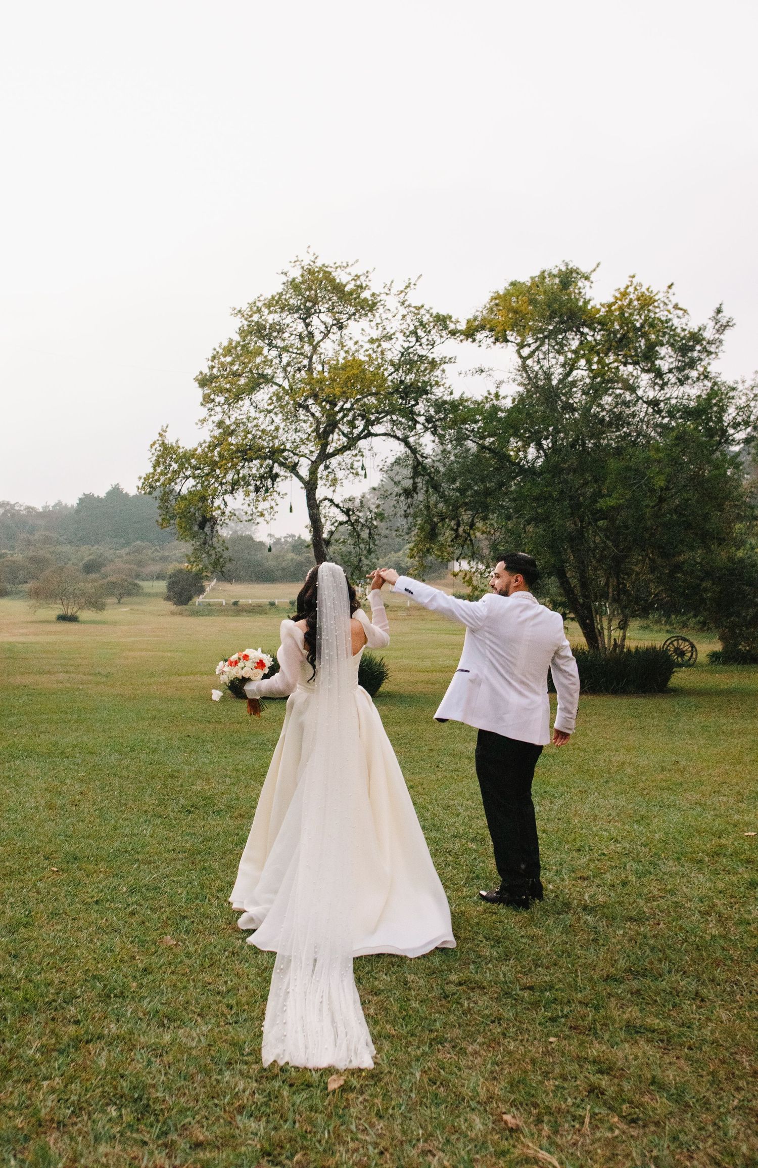 A wedding couple dancing playfully in a grassy field with trees in the background.