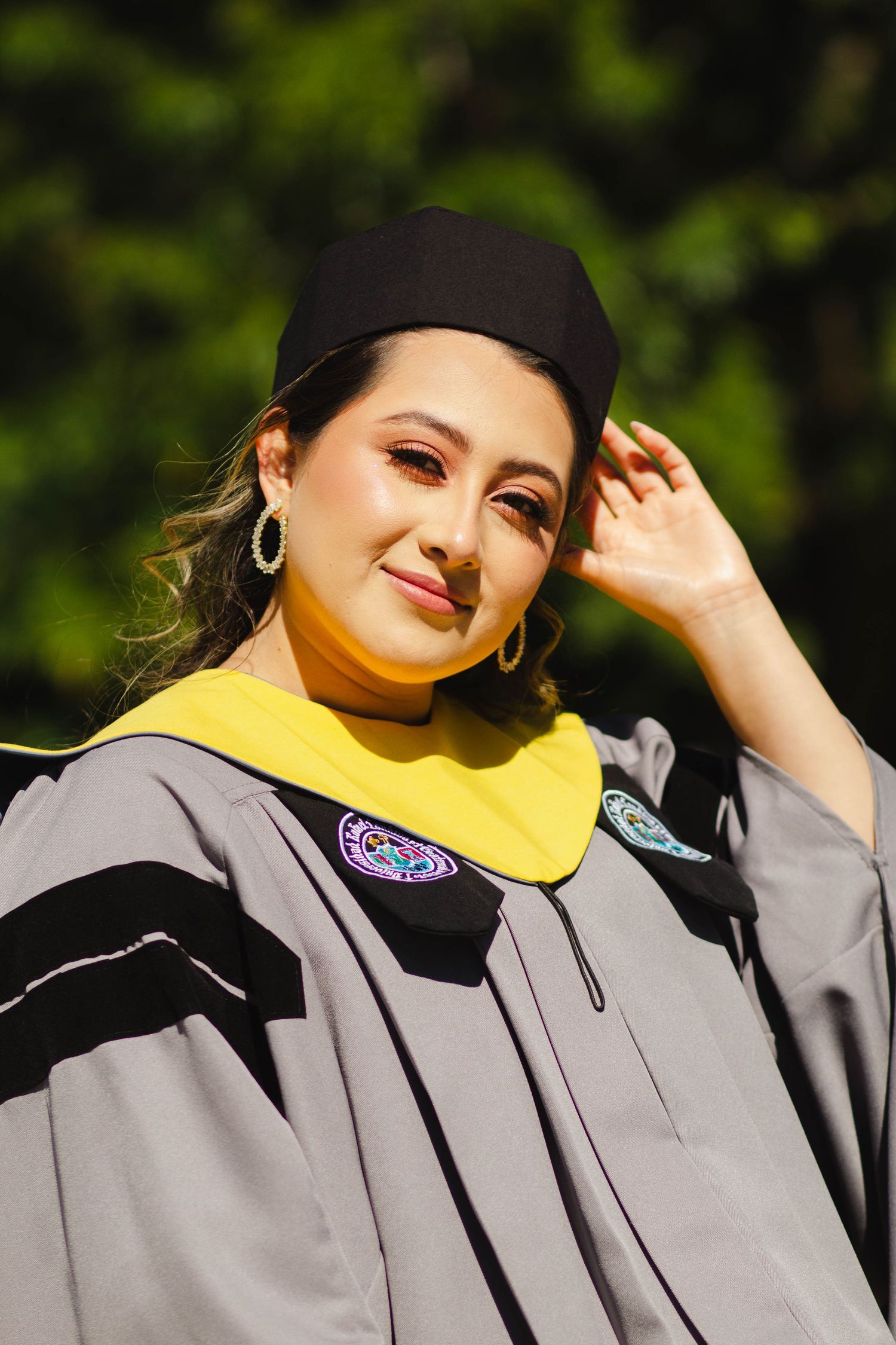 Graduate in academic regalia with yellow stole and black cap poses outdoors on sunny day.