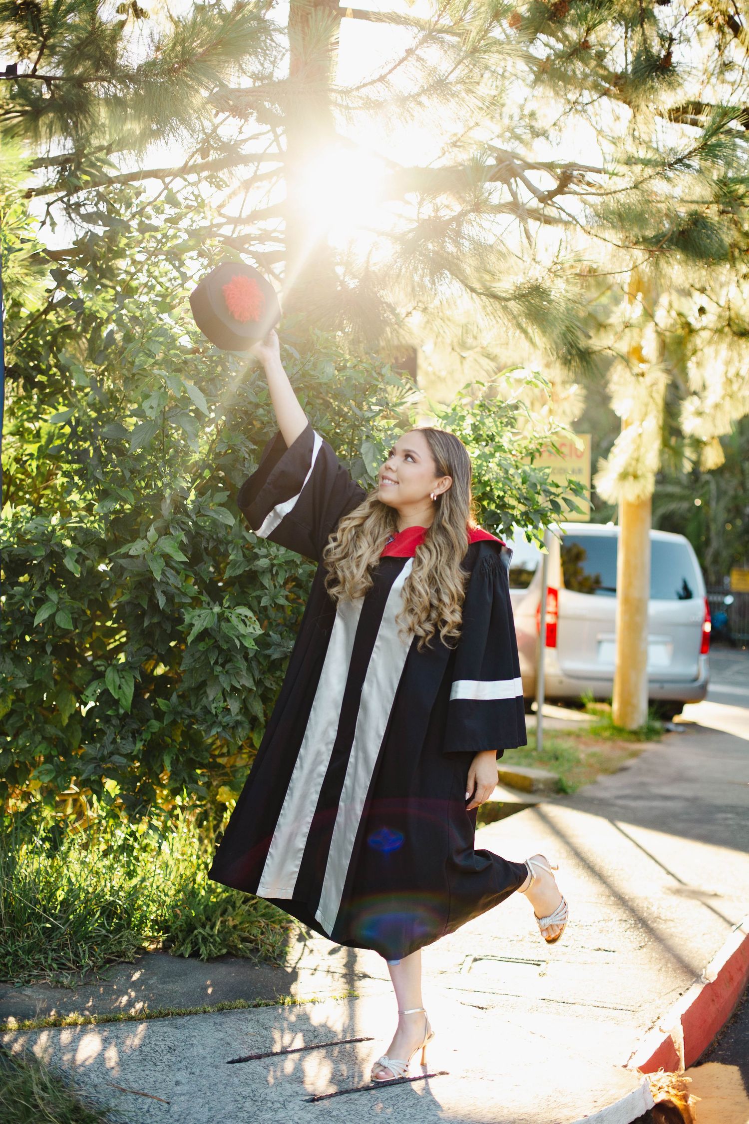 Graduate in black academic regalia holds up cap while posing outdoors in sunlit garden setting.