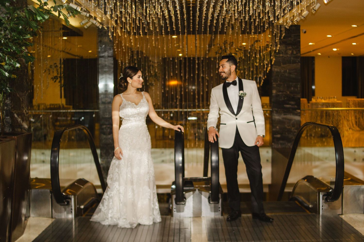 Wedding couple poses elegantly on escalator in luxurious venue with crystal decor.