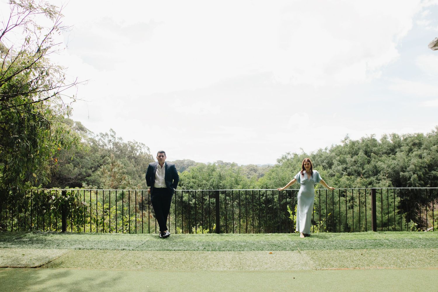 Two people walk playfully down an outdoor pathway bordered by greenery and an iron railing on a sunny day.