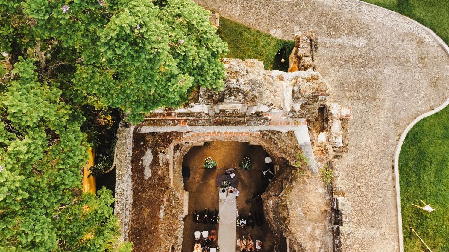 Aerial drone view of ancient stone castle ruins with curved pathway and green trees surrounding the structure.