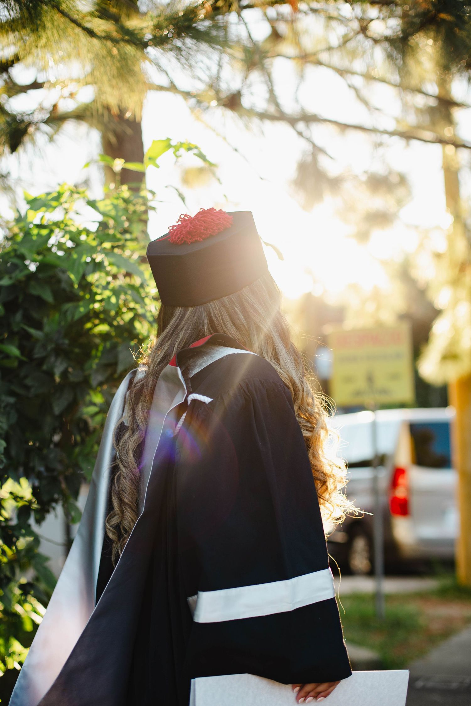 Side view of graduate in black and white academic regalia standing in dappled sunlight.