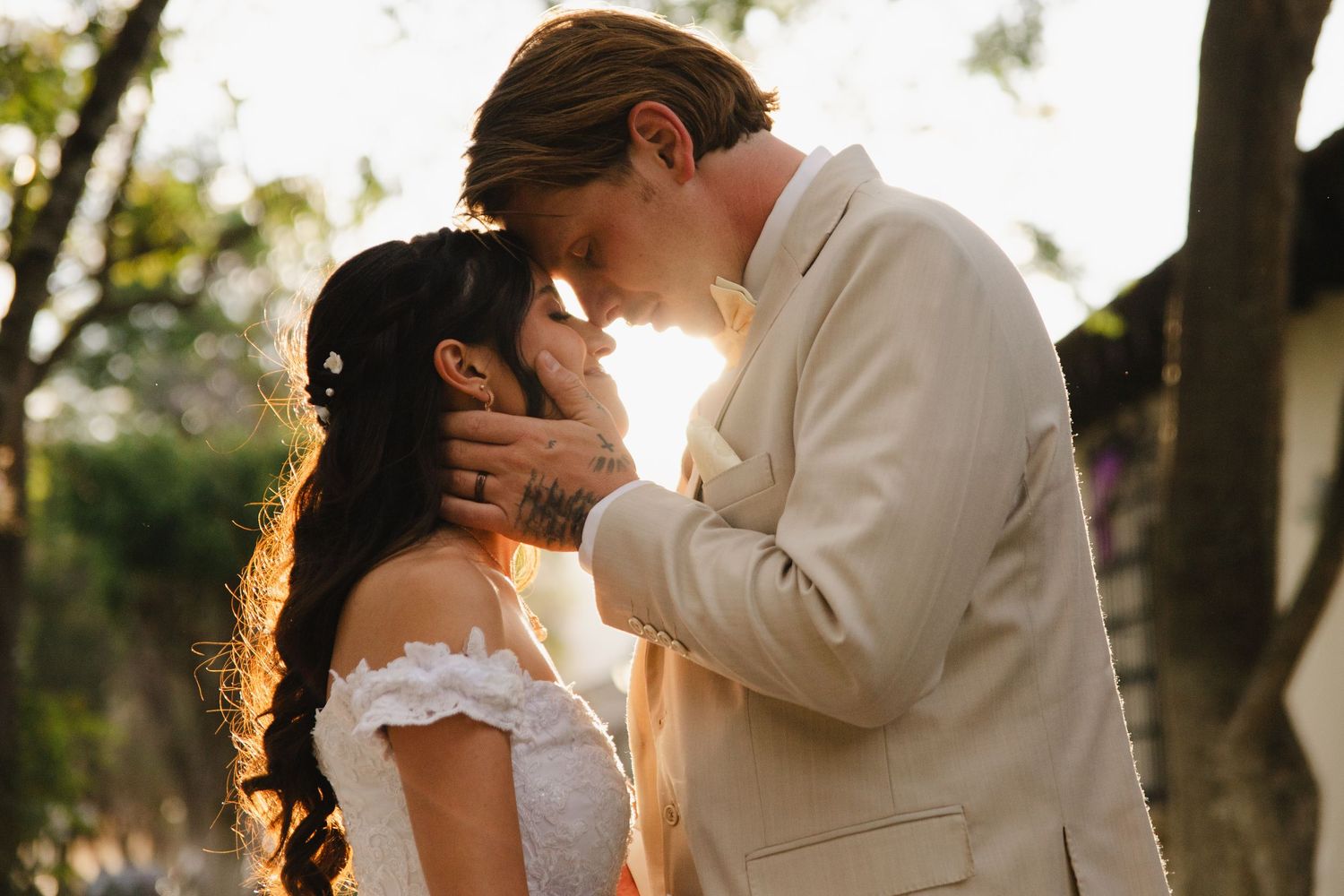 A bride and groom share an intimate moment, touching foreheads outdoors in golden sunlight on their wedding day.
