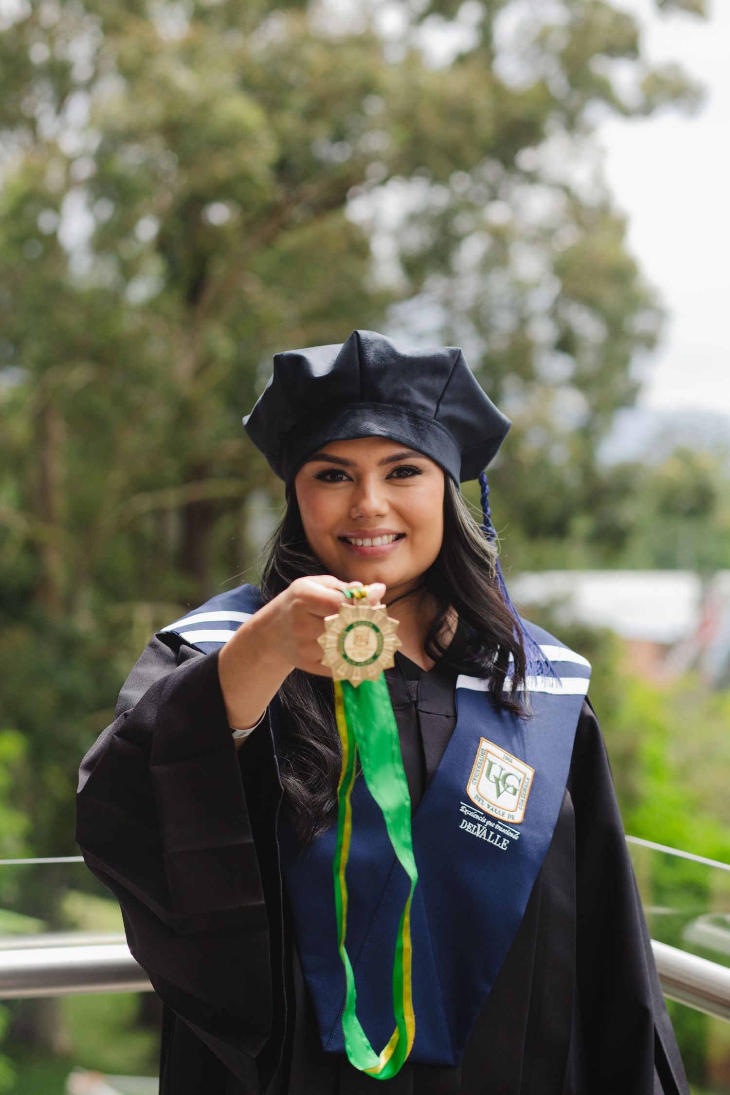 A graduate in black academic regalia holds up a green and gold medallion during an outdoor graduation ceremony.