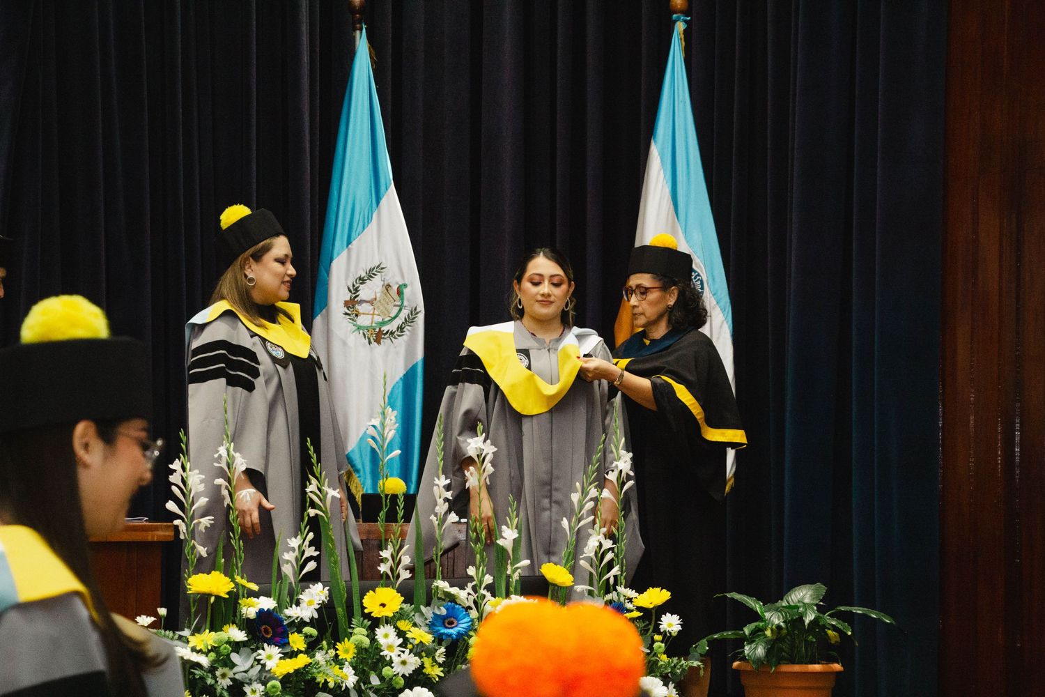 Graduation ceremony with flags and yellow decor featuring graduates in academic regalia on stage.
