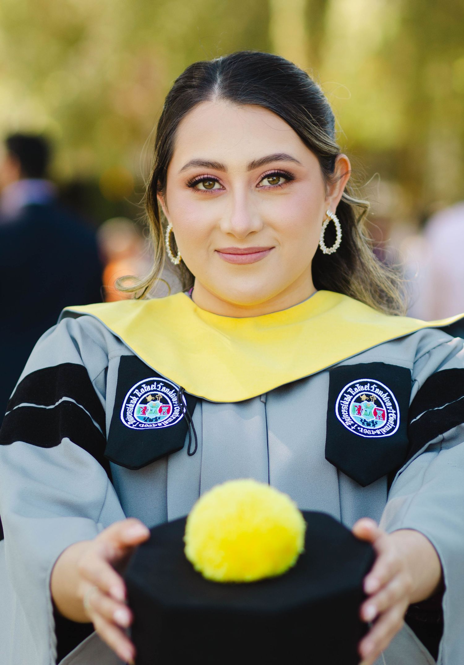 Graduate in academic regalia holds mortarboard against sunny outdoor background.