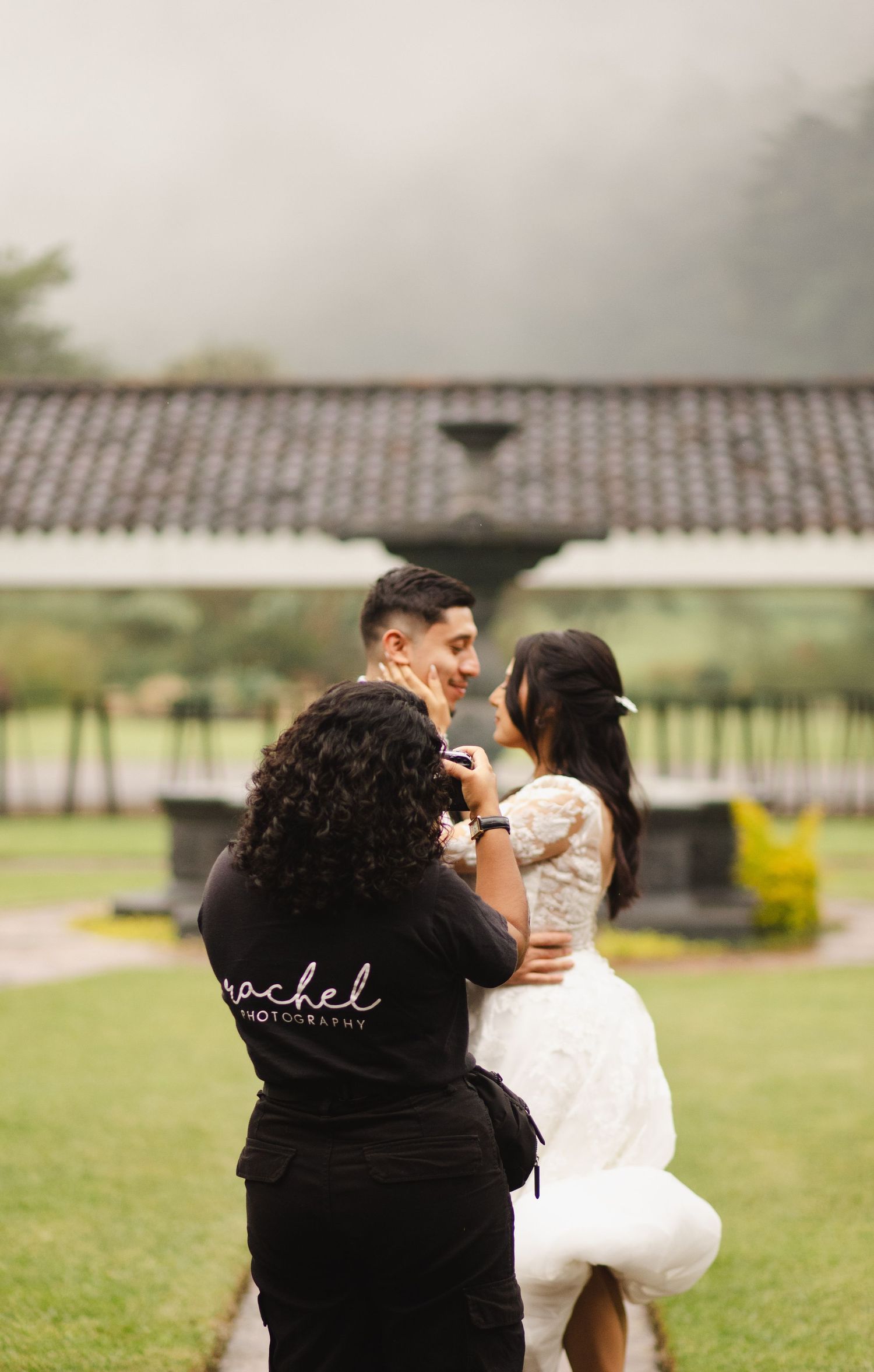 A photographer in black captures images of a couple in a garden setting with Spanish-style architecture.