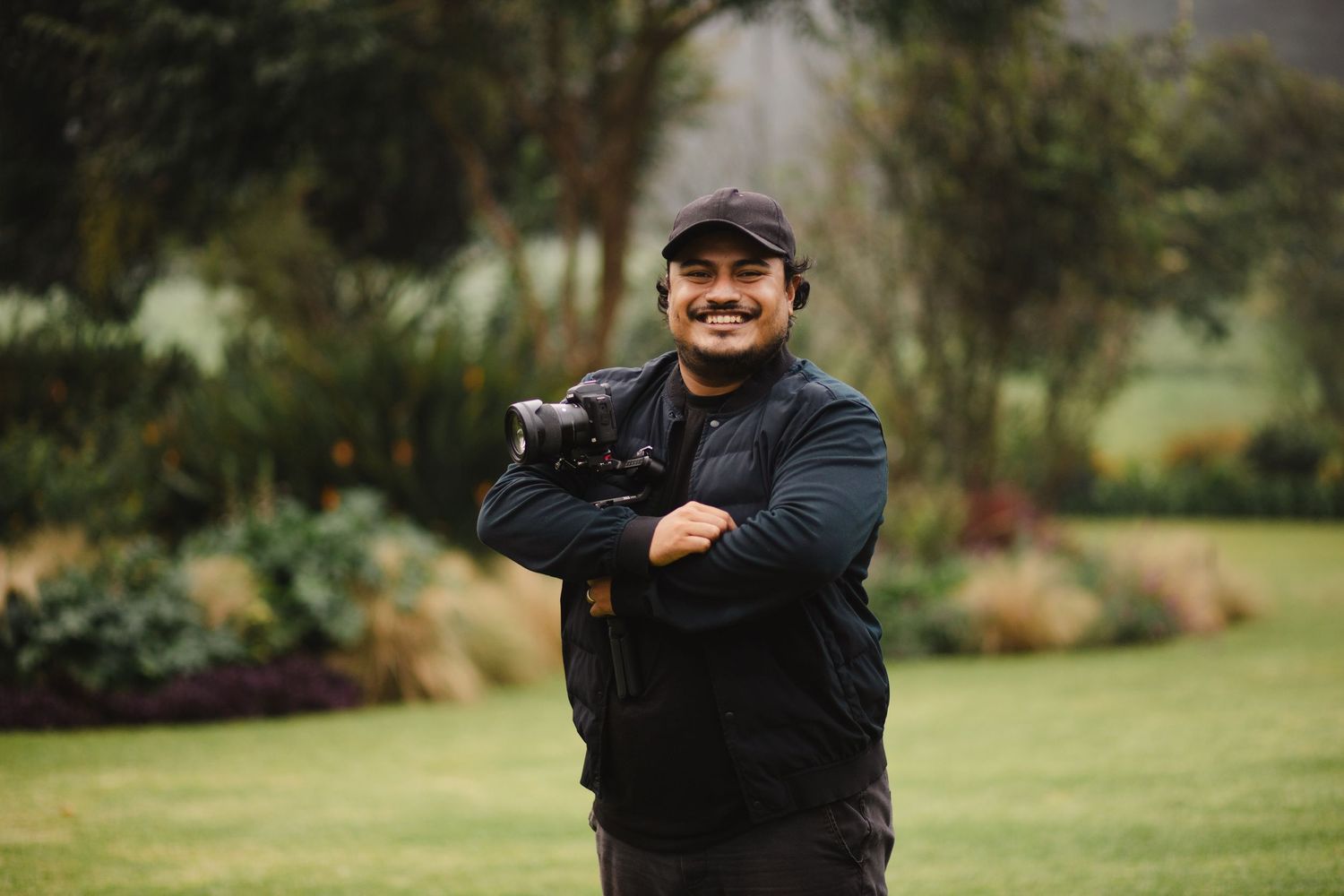 A person in a black jacket and baseball cap stands with crossed arms in front of a lush garden landscape.