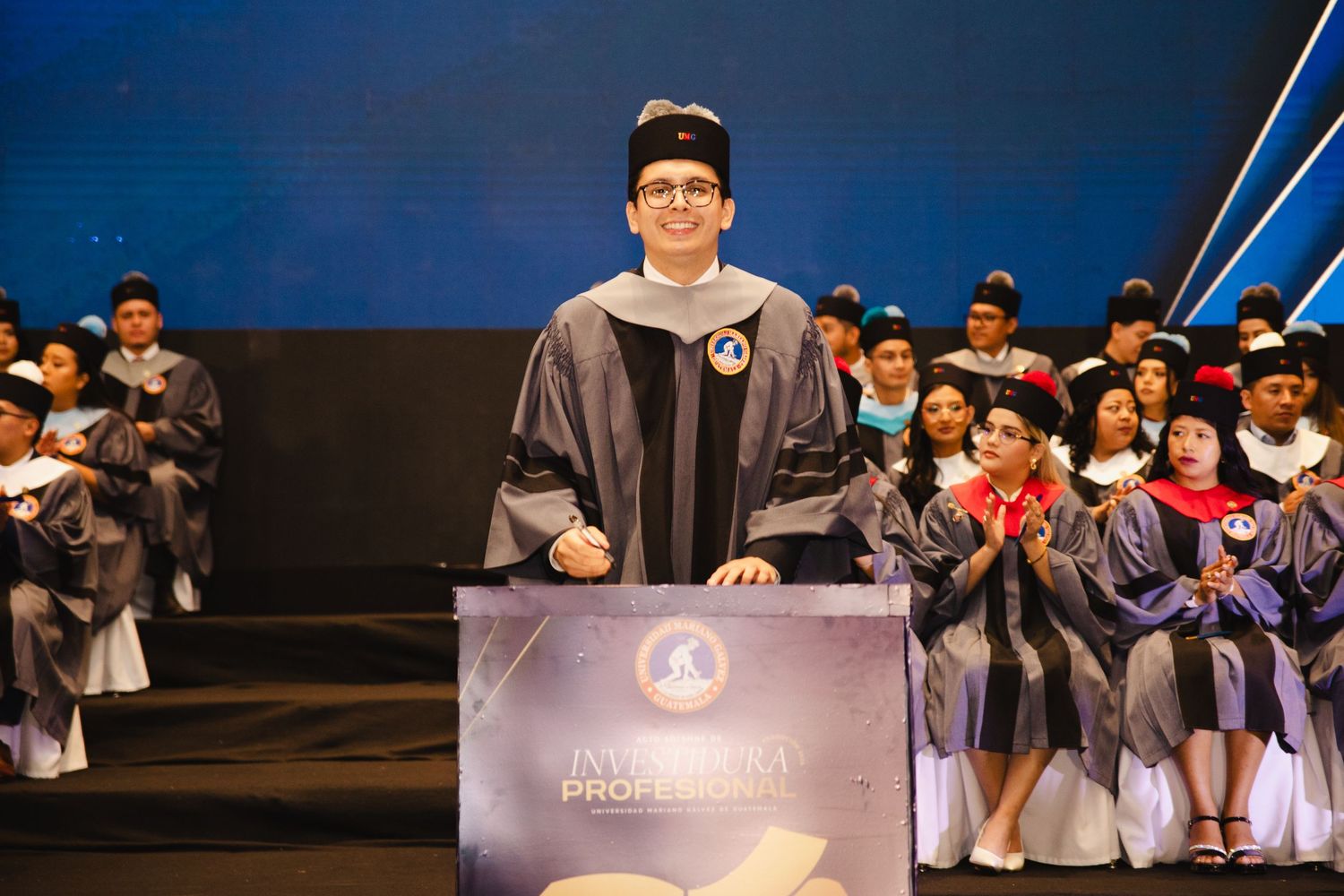 Man in academic regalia stands at podium during university graduation ceremony with graduates seated in background.