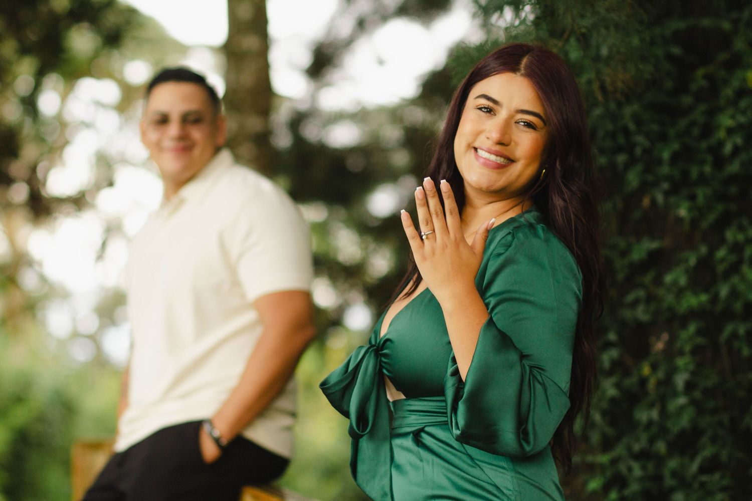 Romantic engagement photo of couple in outdoor setting with woman in green dress showing engagement ring.