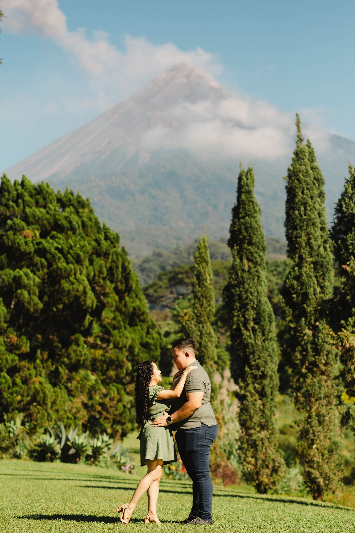 A couple poses romantically on a green lawn with a majestic volcano visible in the background.